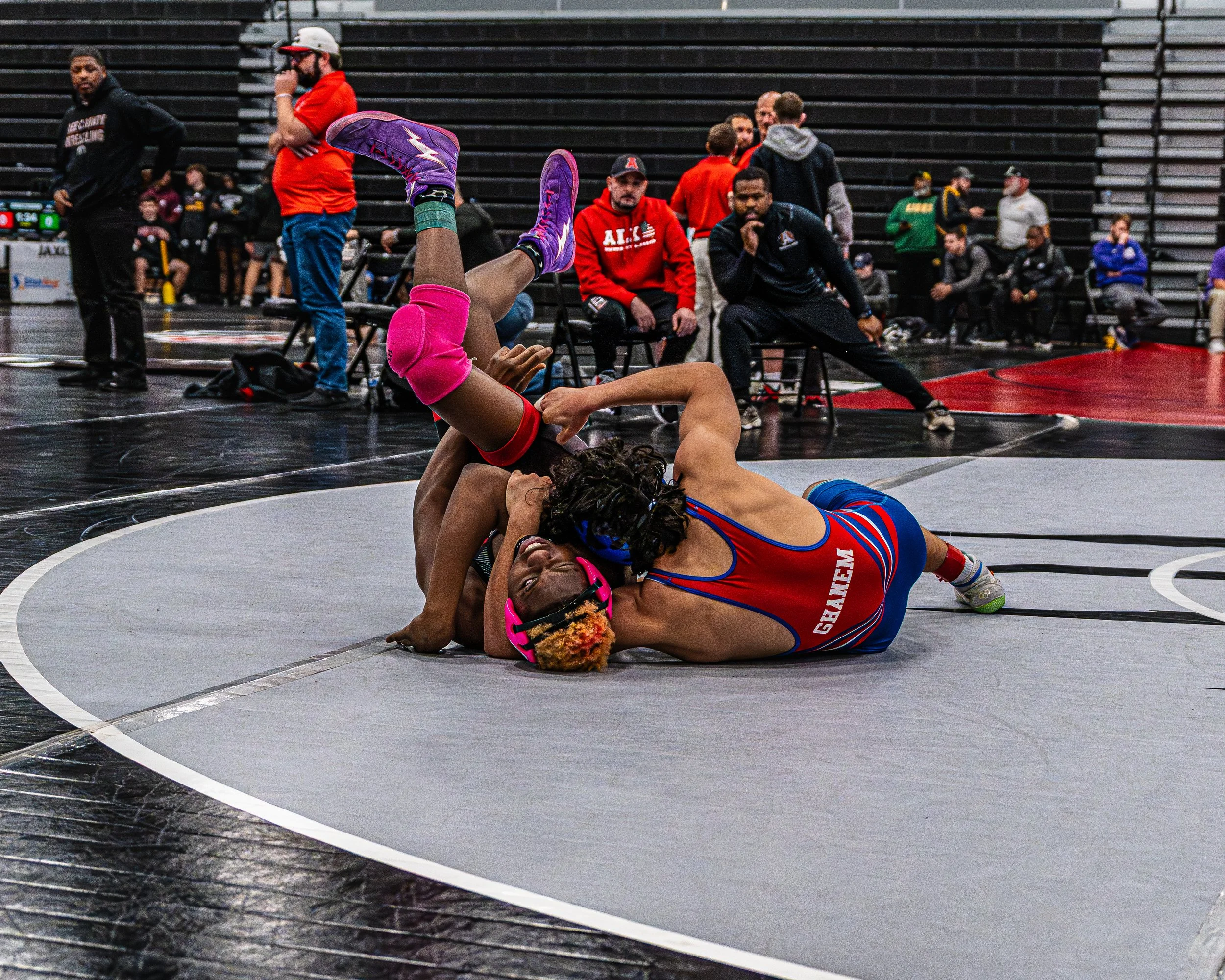 Two female wrestlers are engaged in a match on a wrestling mat, with one wrestler holding the other down while wearing a red and blue singlet and the other in pink gear. Several spectators and officials observe the match in a gymnasium.