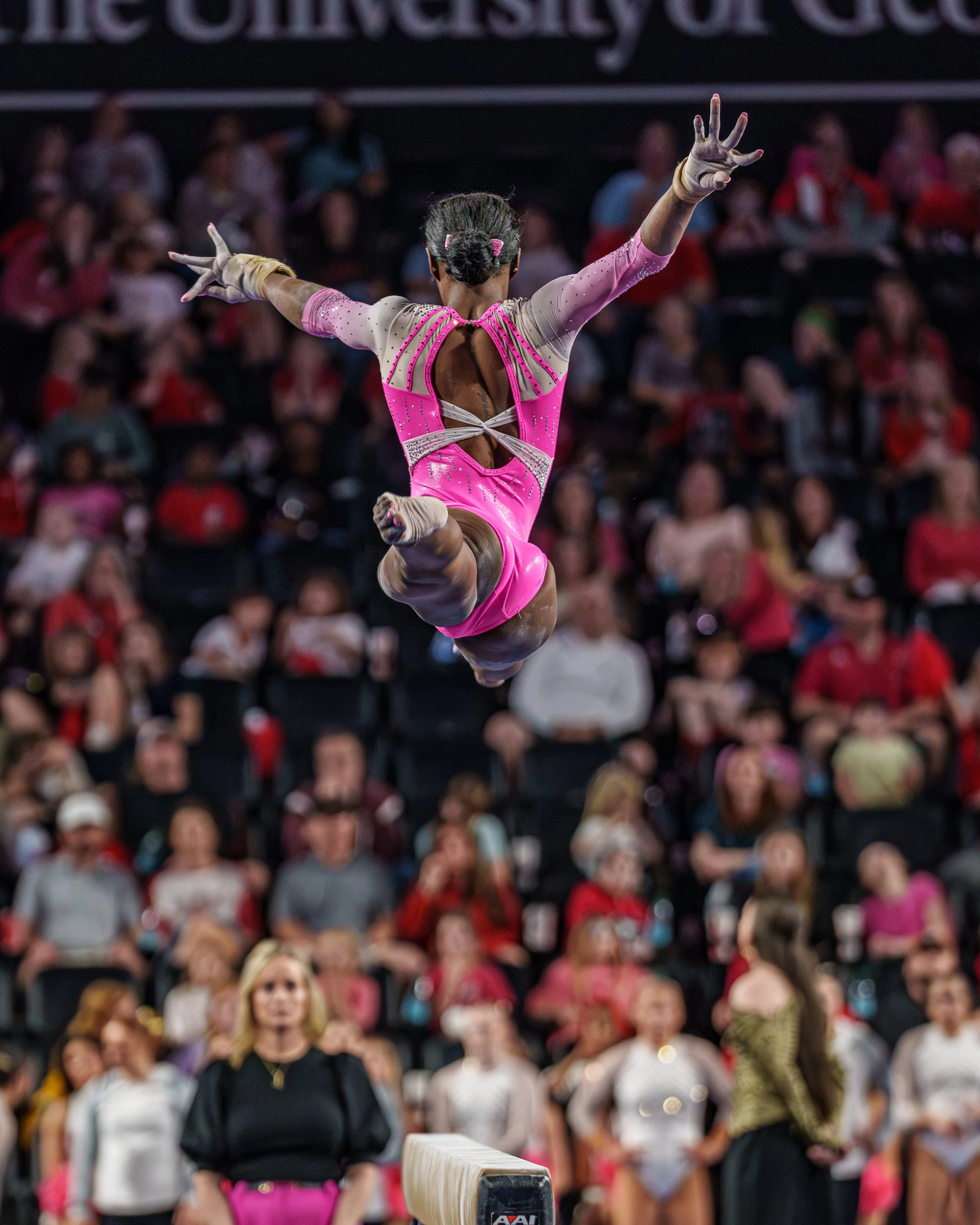 A female gymnast in a pink and beige leotard performing a tumbling flip during a gymnastics competition, with an audience watching in the background.