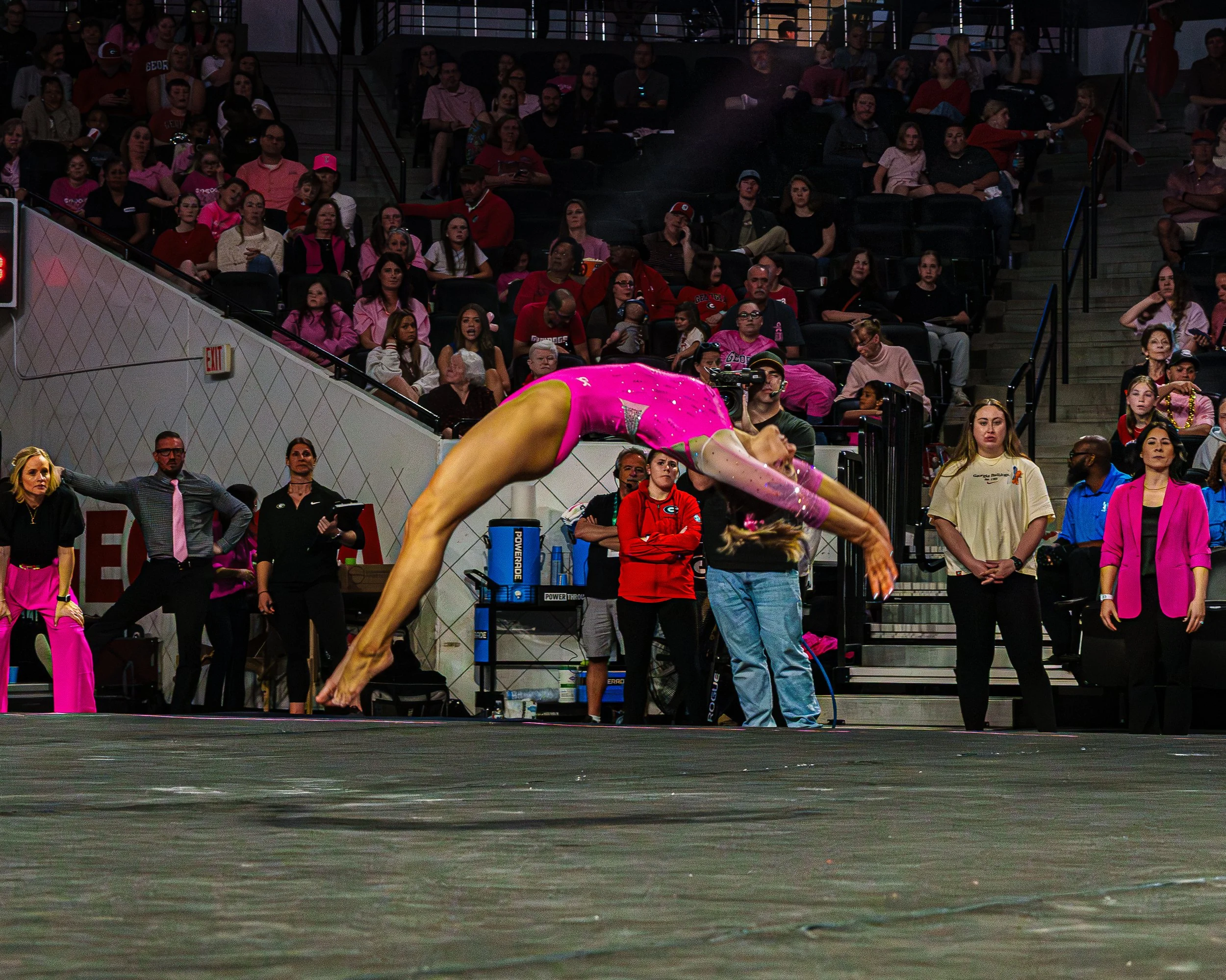 A female gymnast in a pink leotard with sparkly details performing a backbend on a gymnastics mat during a competition, with spectators watching from the stands.