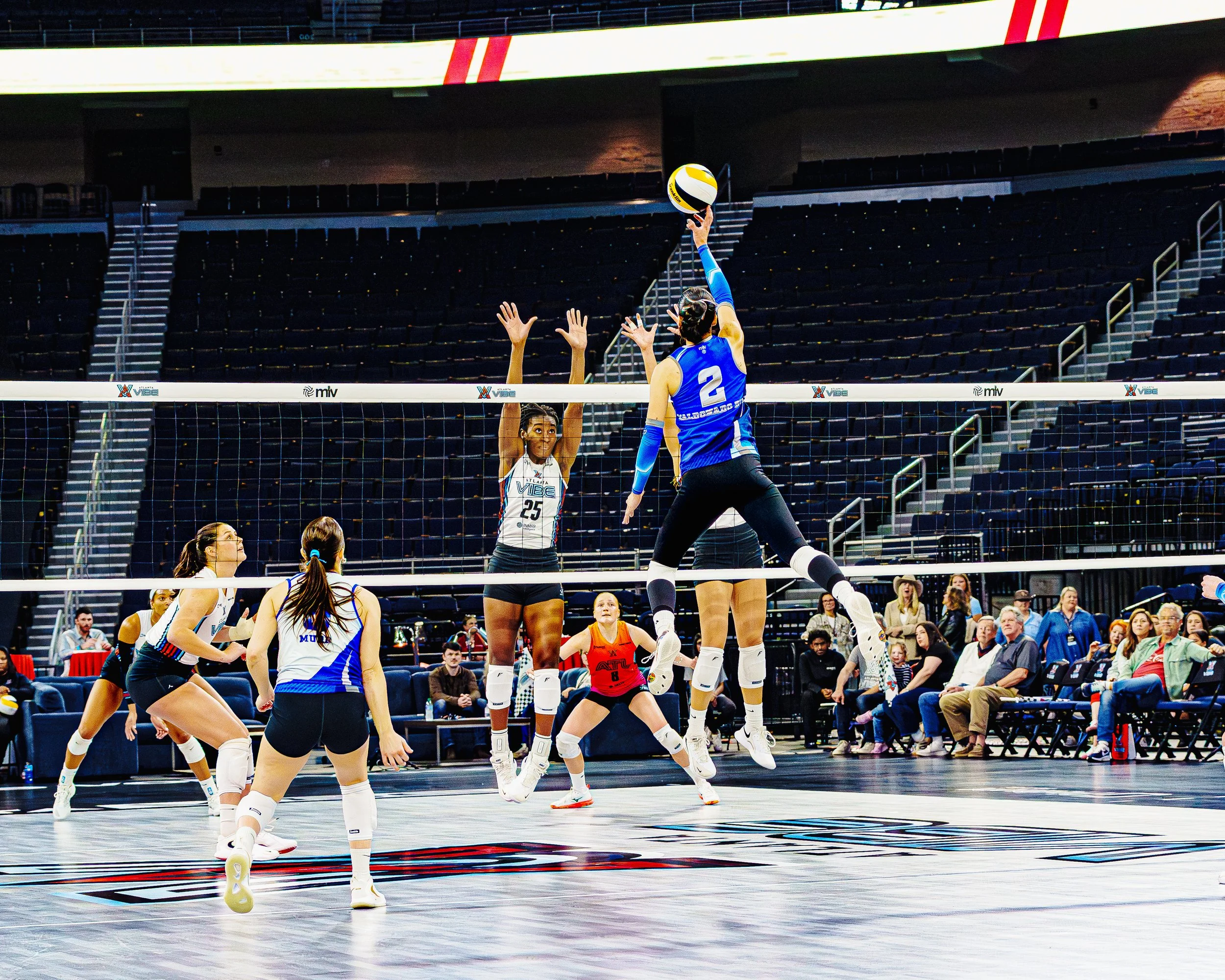 Women playing volleyball indoors, with one player jumping to spike the ball while others prepare to block or receive.