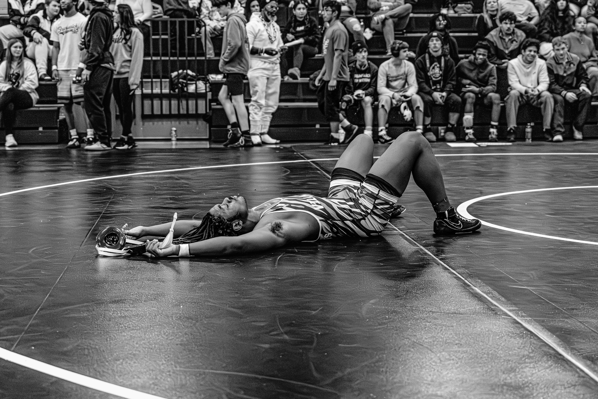 A female wrestler lying on her back on a wrestling mat, holding a trophy in one hand and a medal in the other, with spectators watching in the background.