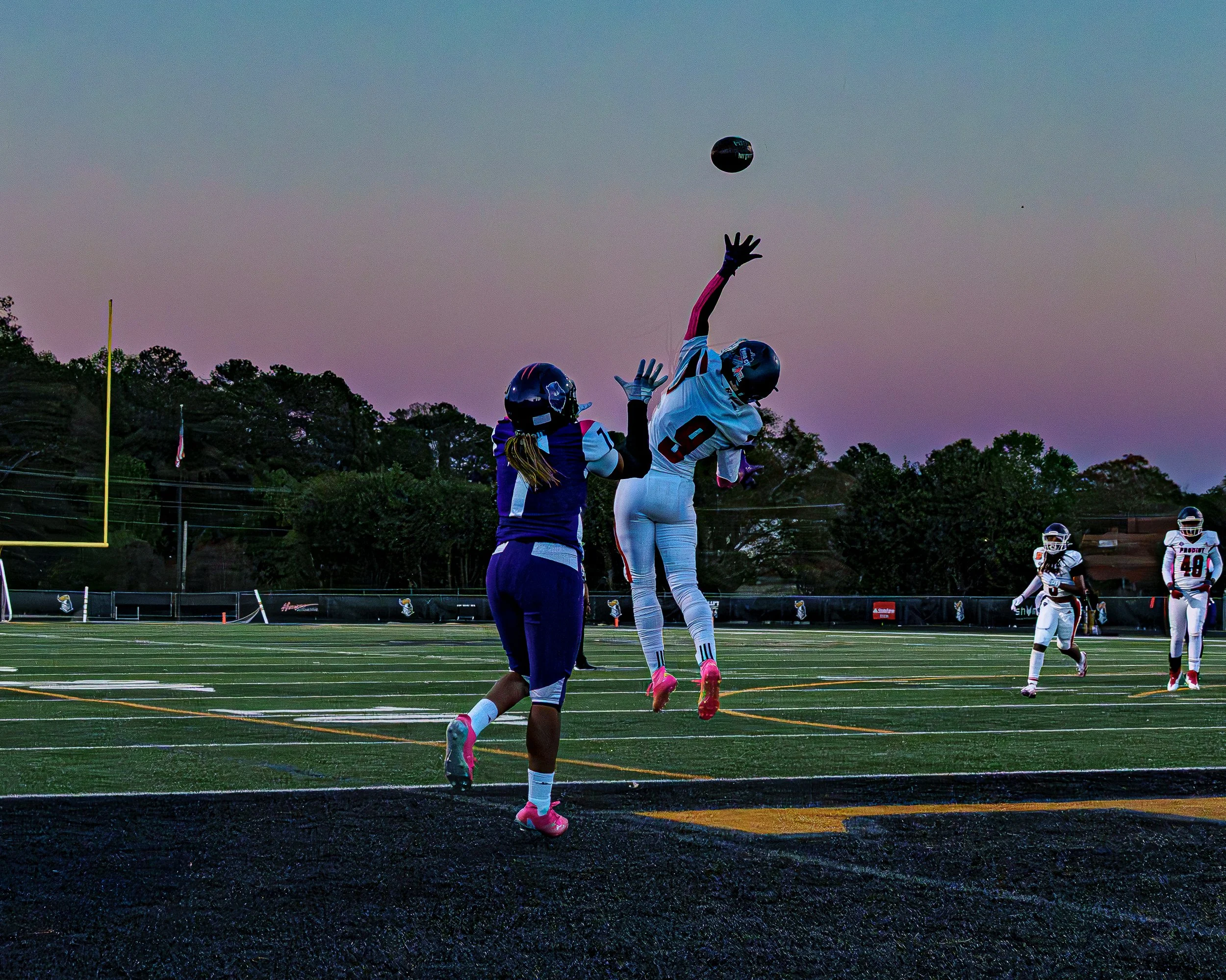 Two football players mid-play on a field during dusk, one in a white uniform jumping to catch the ball, the other in purple trying to block.