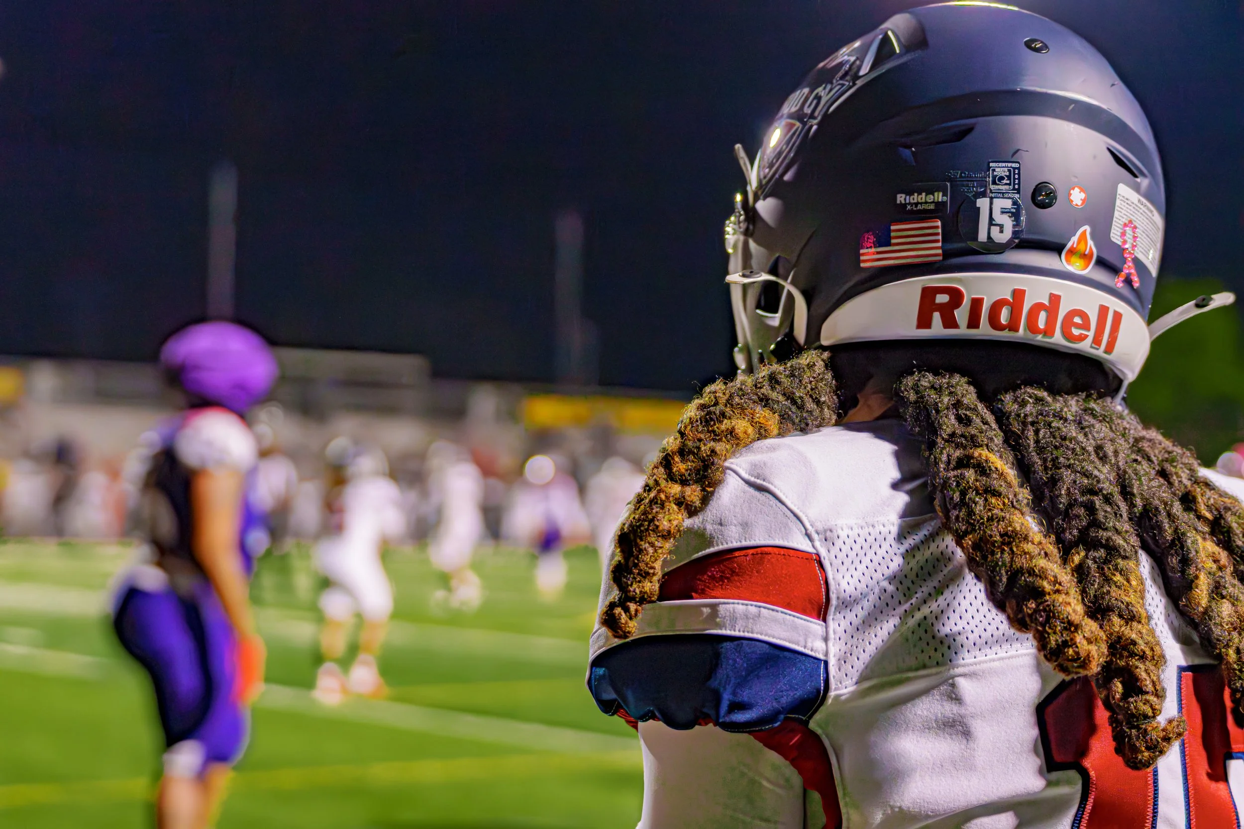 Close-up of a football player with long dreadlocks wearing a black helmet and white uniform during a game at night, with teammates and opponents on the field in the background.