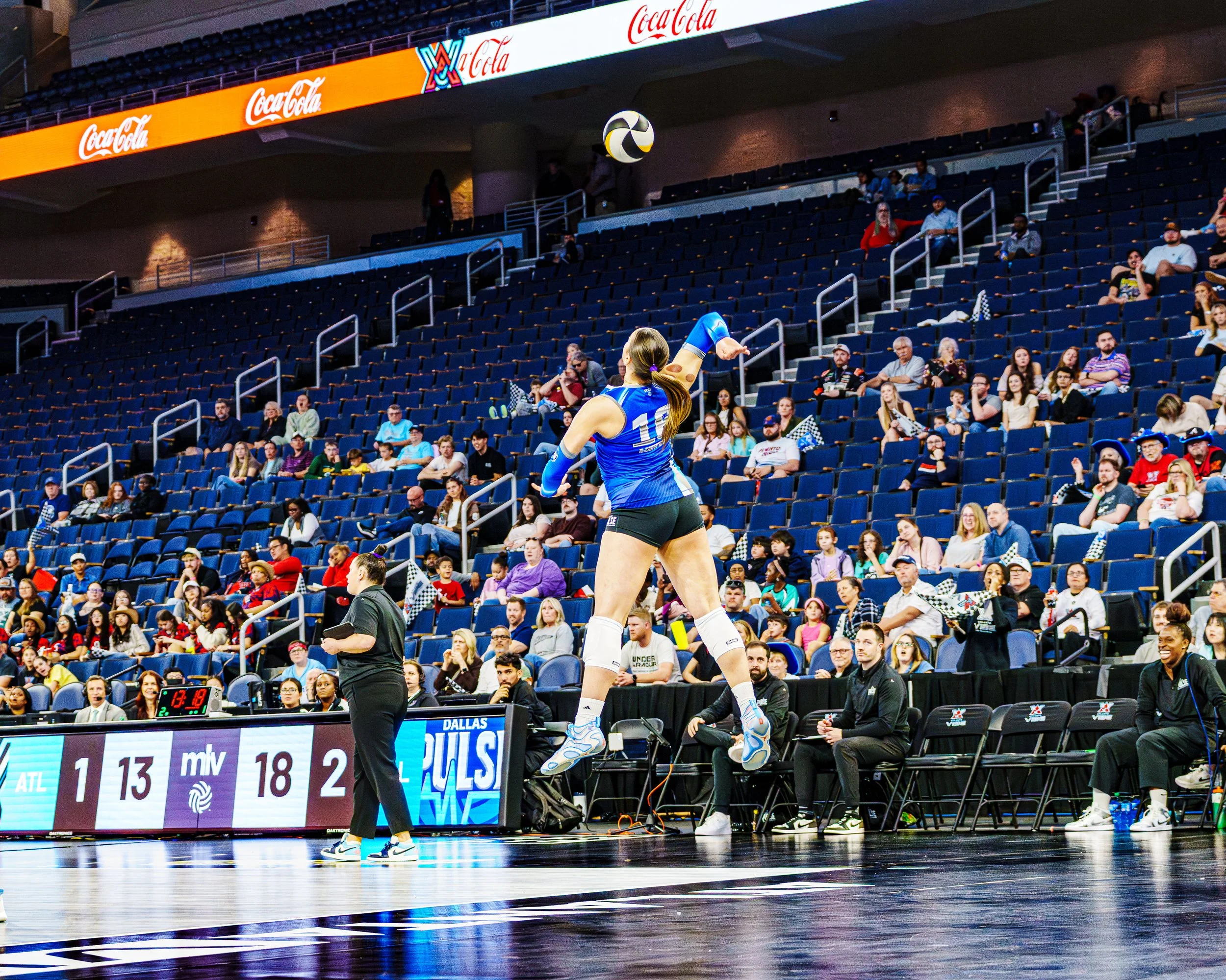 A volleyball player in a blue uniform is jumping to hit the volleyball in a stadium with spectators and officials, with scoreboard and advertisements visible.
