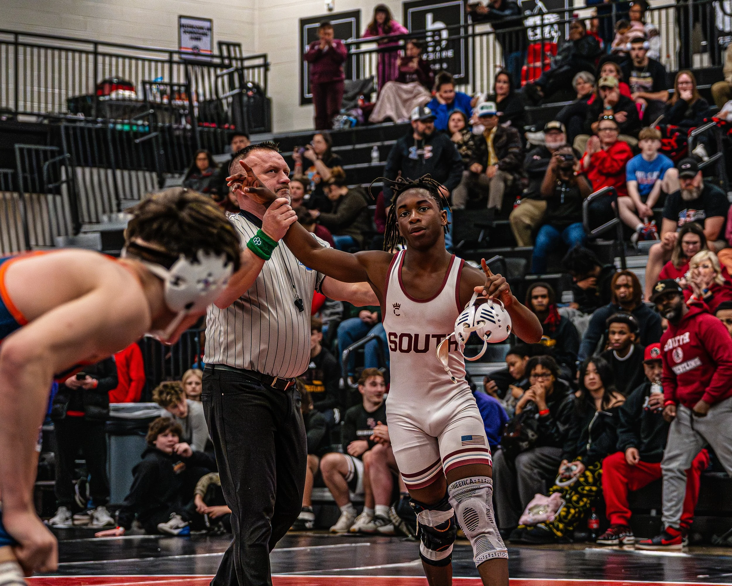 A female wrestler in a white singlet with maroon accents, holding a helmet, is being awarded a victory gesture by a referee in a gymnasium. A male wrestler with a mask is bent over nearby, and an audience sits in the background watching.