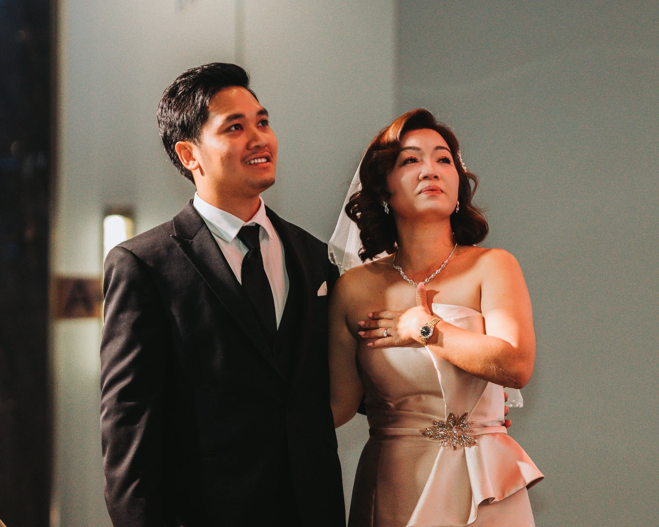A moment during a wedding ceremony showing a bride and groom standing together, with the bride's hand over her heart, in a room with simple decor.