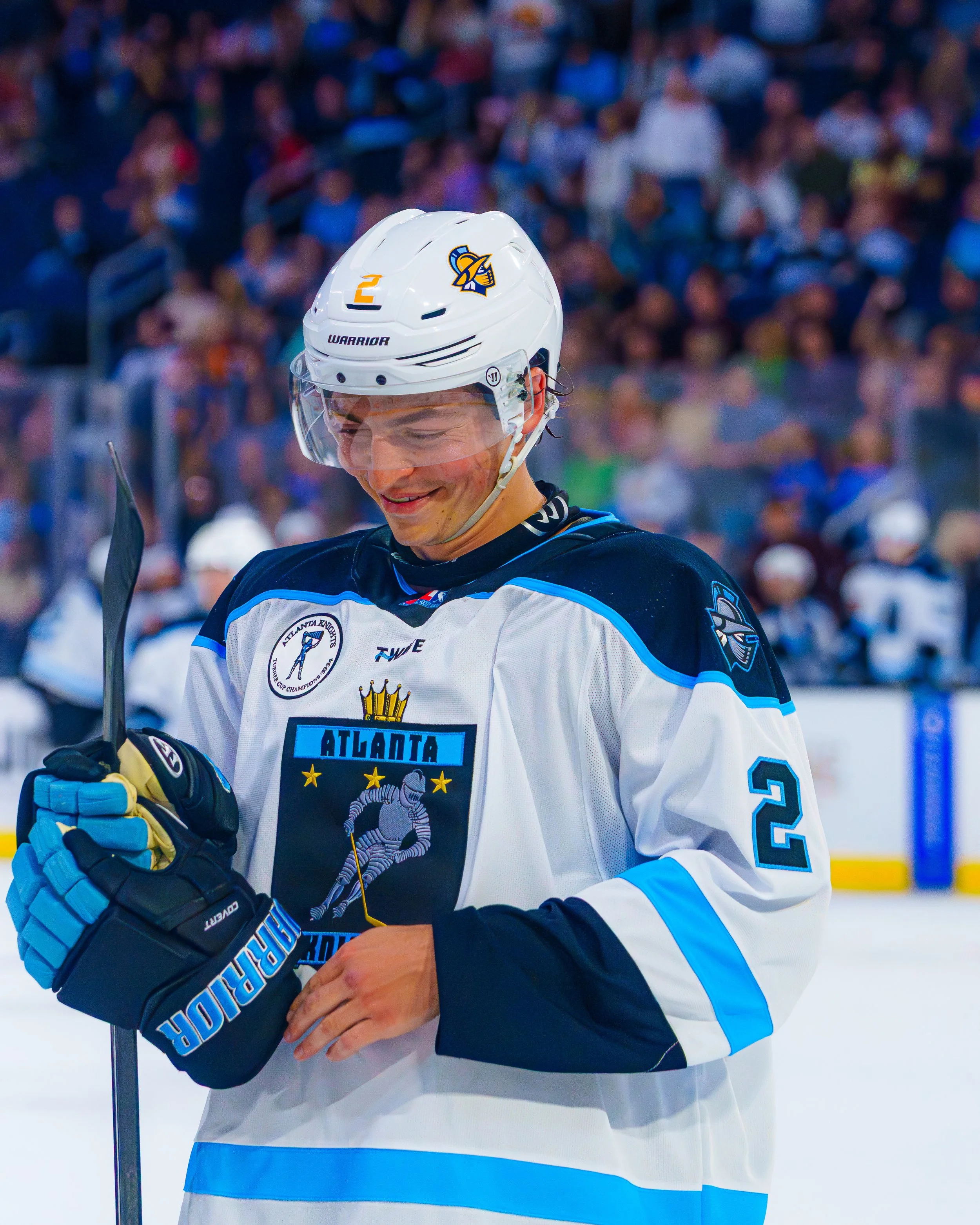 A hockey player in a white jersey with blue accents, wearing a helmet with a team logo, smiling and holding a hockey stick on the ice rink.