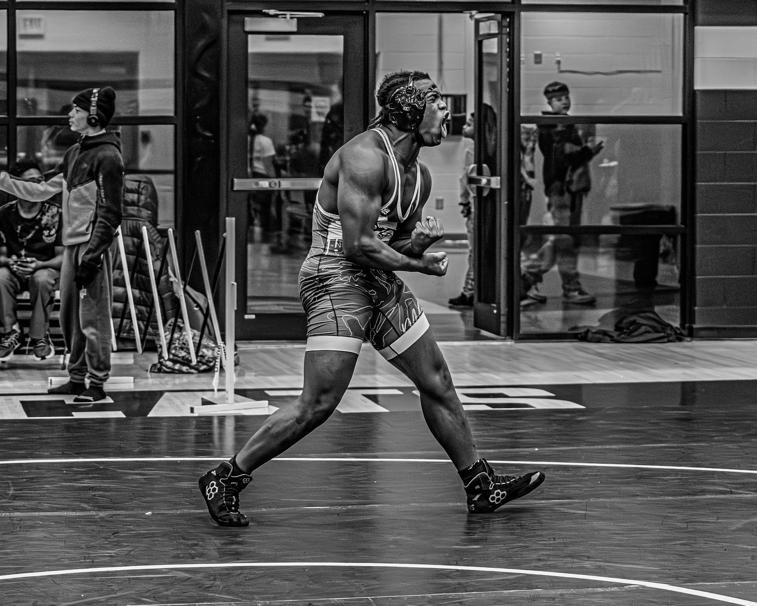 A male wrestler in a singlet celebrates with a loud yell and a clenched fist during a match at an indoor gymnasium. Spectators and coaches are visible in the background through glass doors.