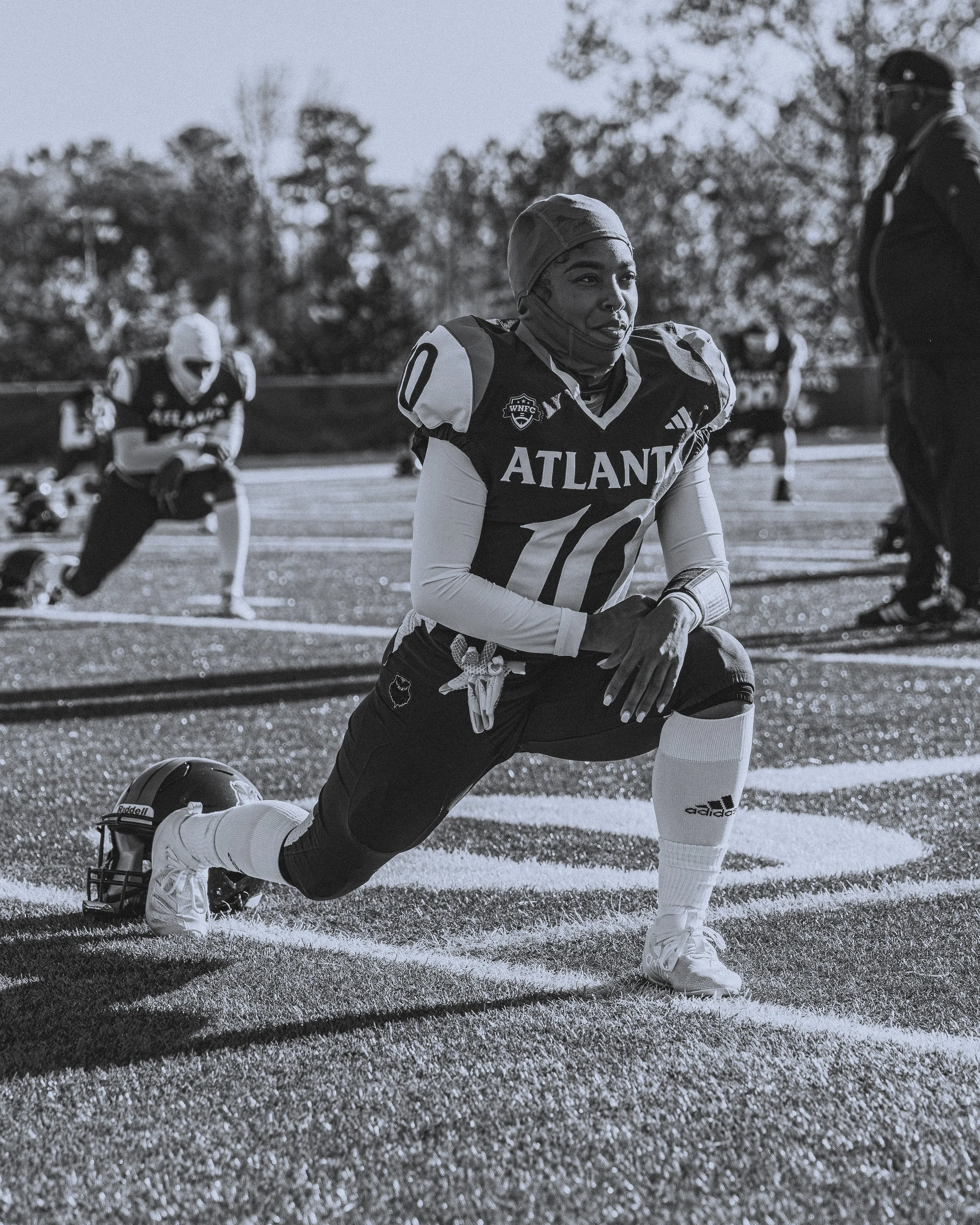 A female football player in uniform kneeling on the field during a game, with other players in the background. She is wearing a jersey with the number 10 and the team name ATLANTA.
