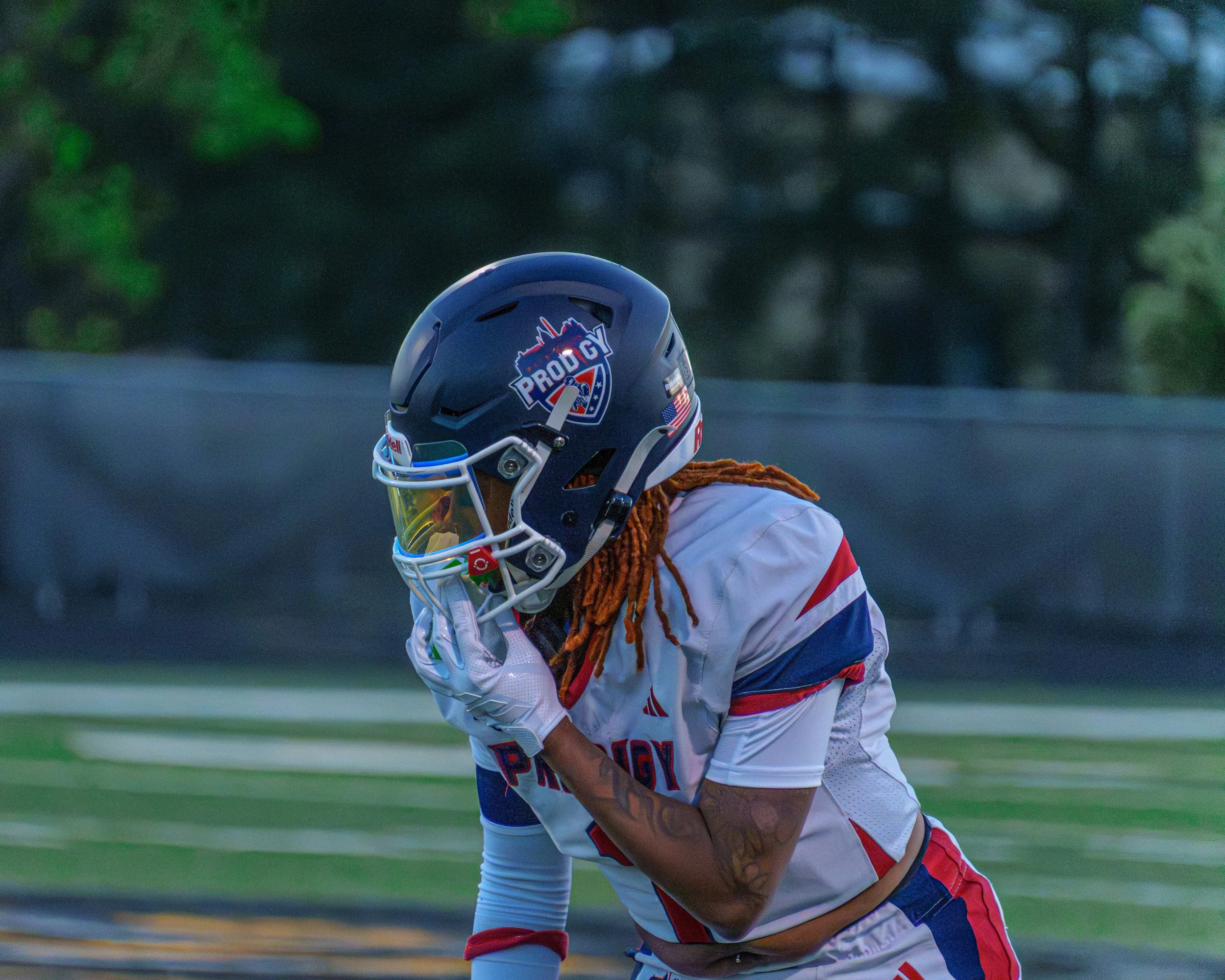 A football player with a helmet and uniform on the field, wearing a helmet with a logo that says 'PRODIGY' and a face shield, appearing to be in deep thought or resting after play.