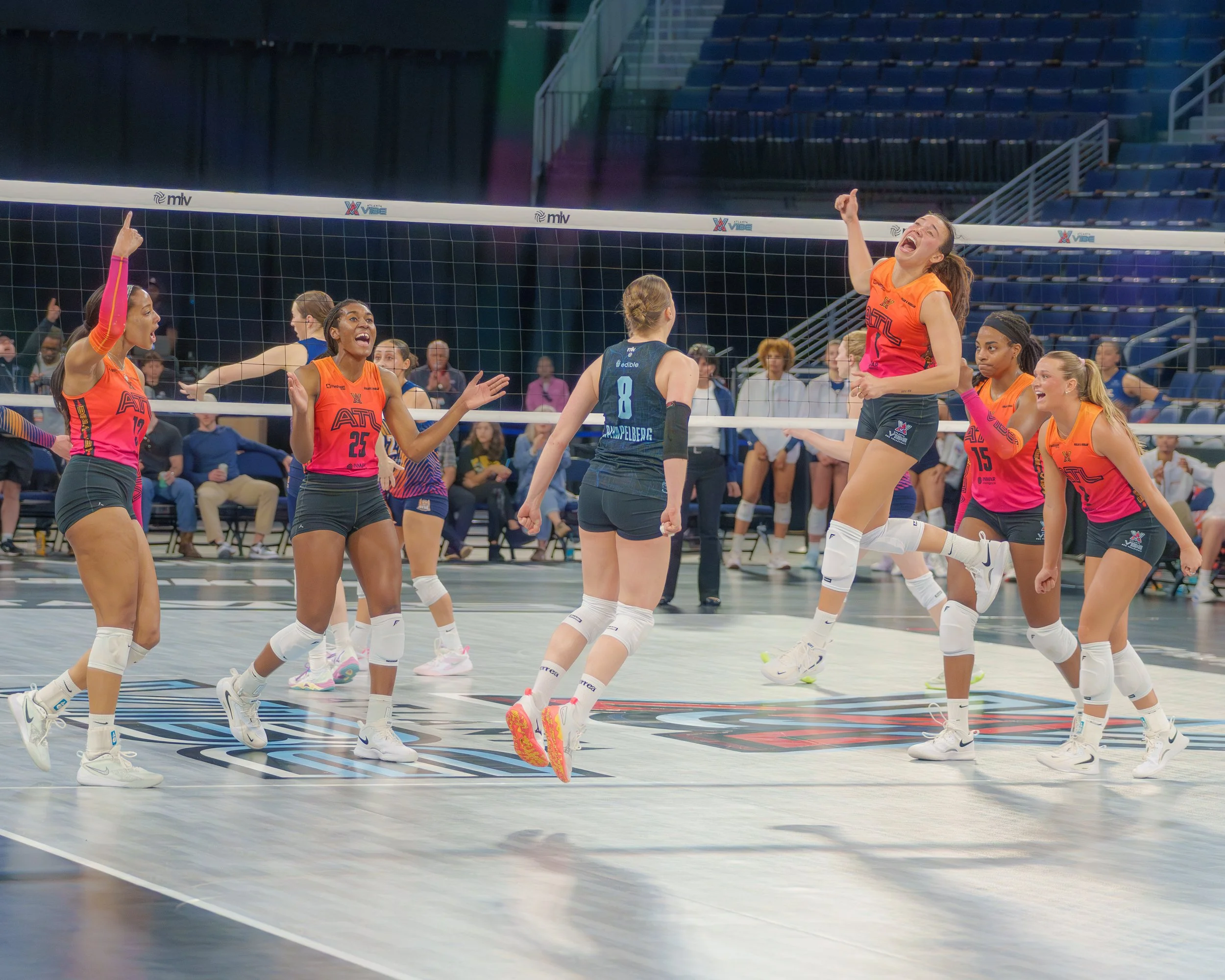 Six volleyball players in colorful uniforms celebrating on the court during a match, with spectators in the background.