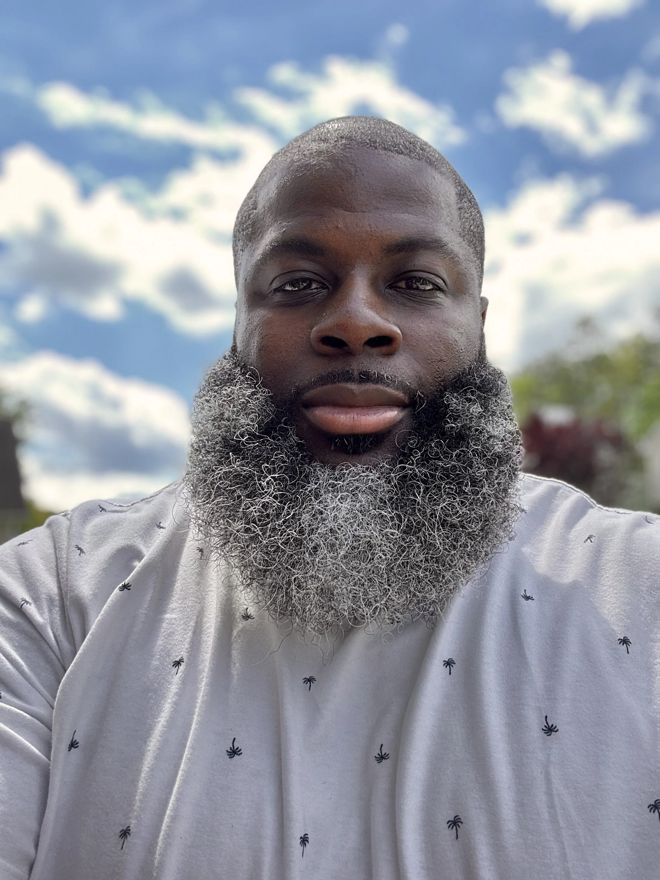 A man with a dark complexion and a full, graying beard takes a selfie outdoors under a partly cloudy sky, with trees in the background.