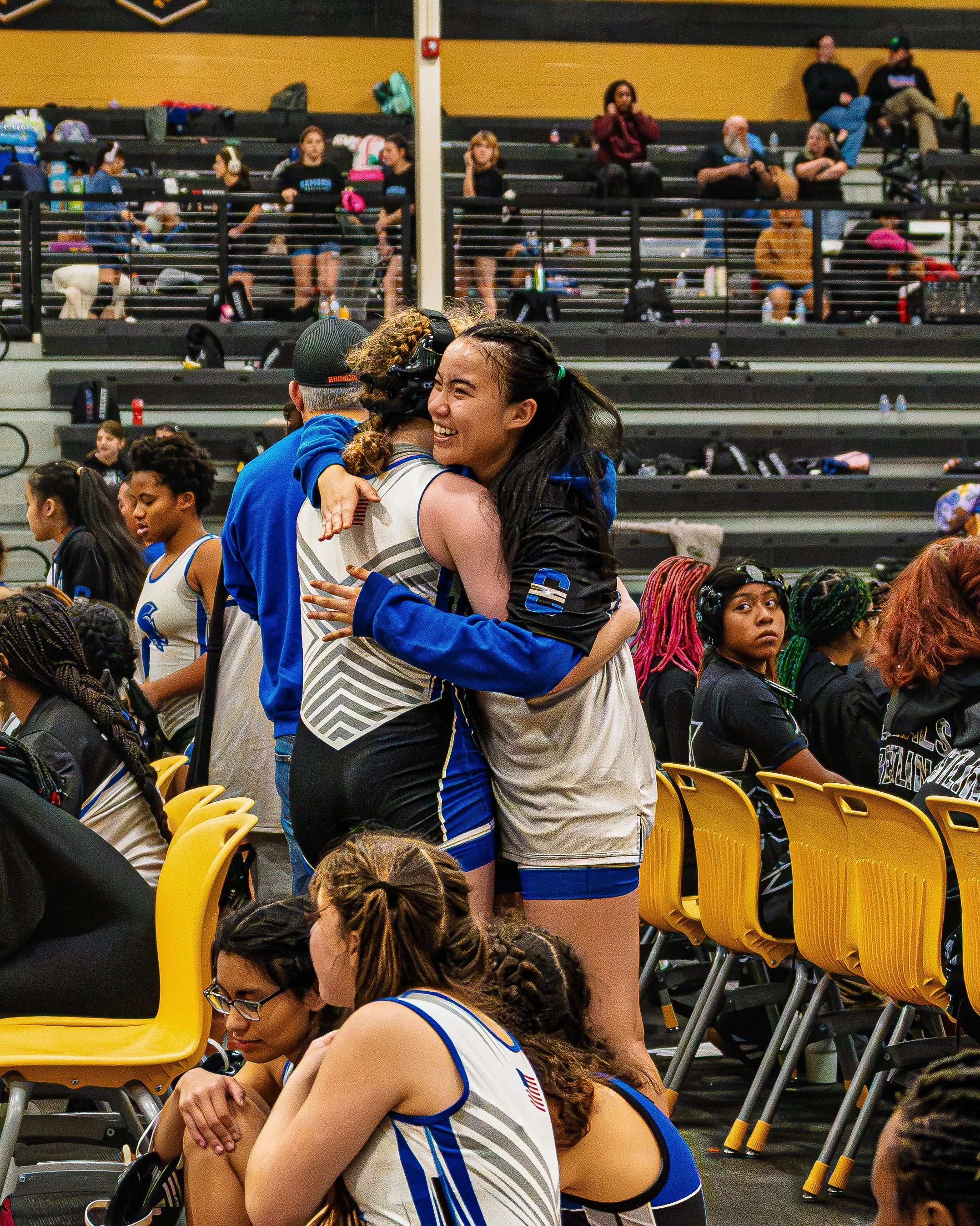 Two female volleyball players embrace in a celebratory hug after a game, surrounded by teammates and spectators in a gymnasium.