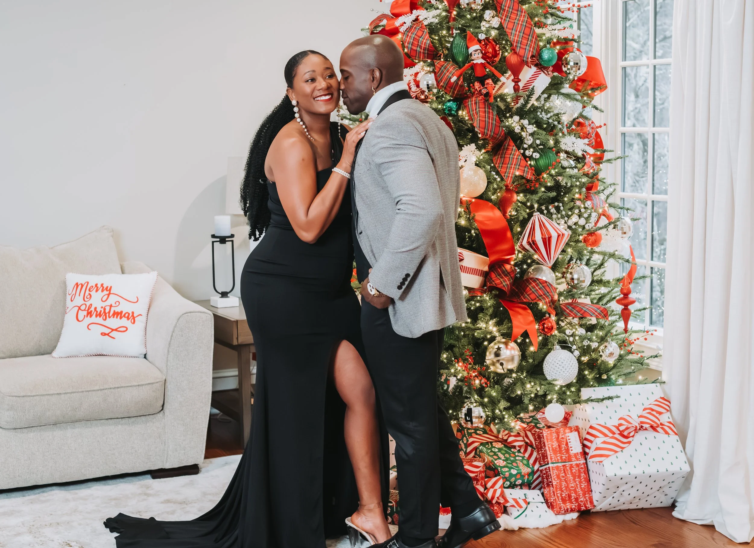 A couple dressed elegantly standing near a decorated Christmas tree with presents underneath in a cozy, well-lit room. The woman is wearing a black evening gown with a high slit, and the man is in a light gray blazer and dark trousers. A pillow with 