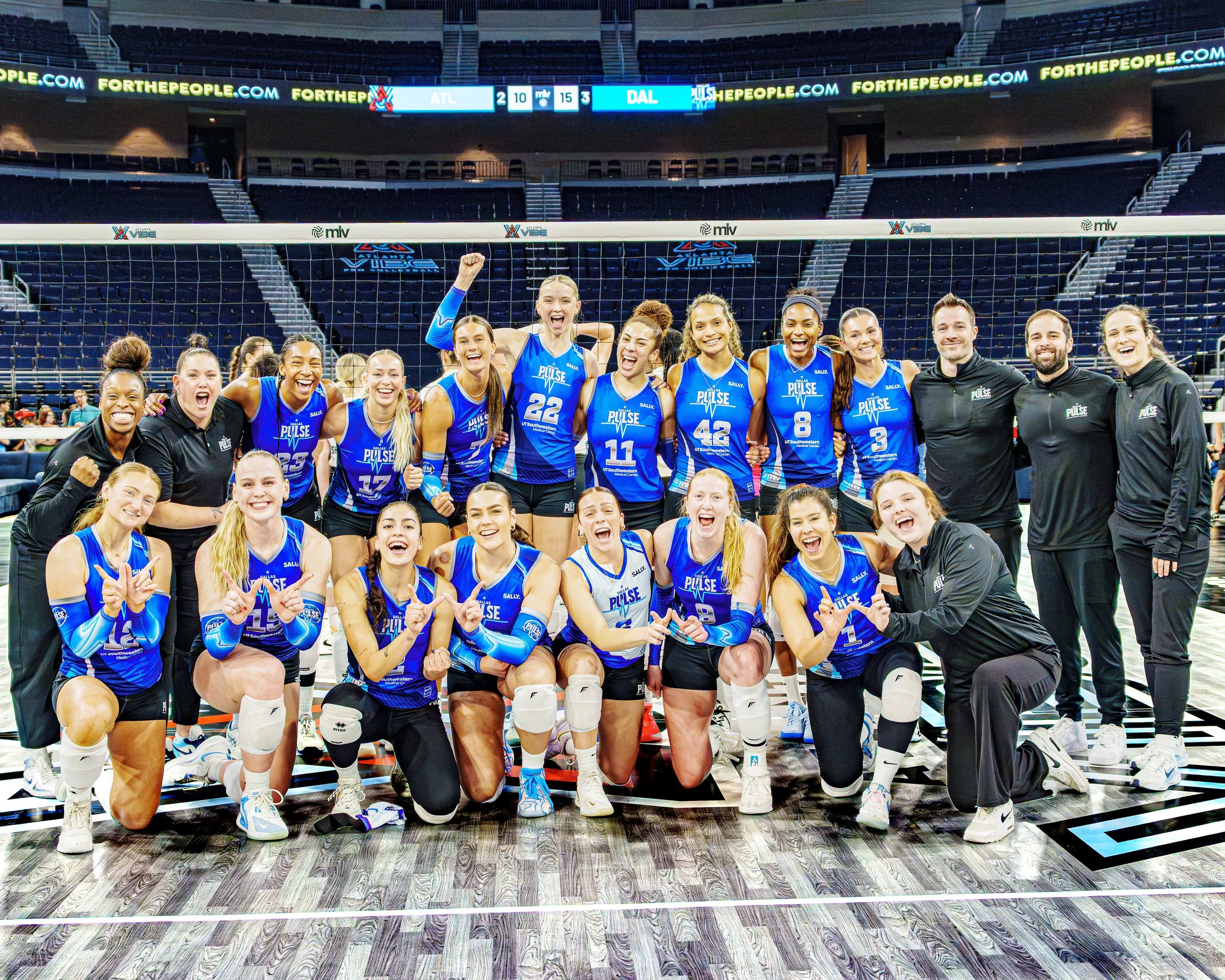 A group of female volleyball players and coaches celebrating on the court after a match, wearing blue uniforms with the word 'Pulse' on them, inside an indoor sports arena.