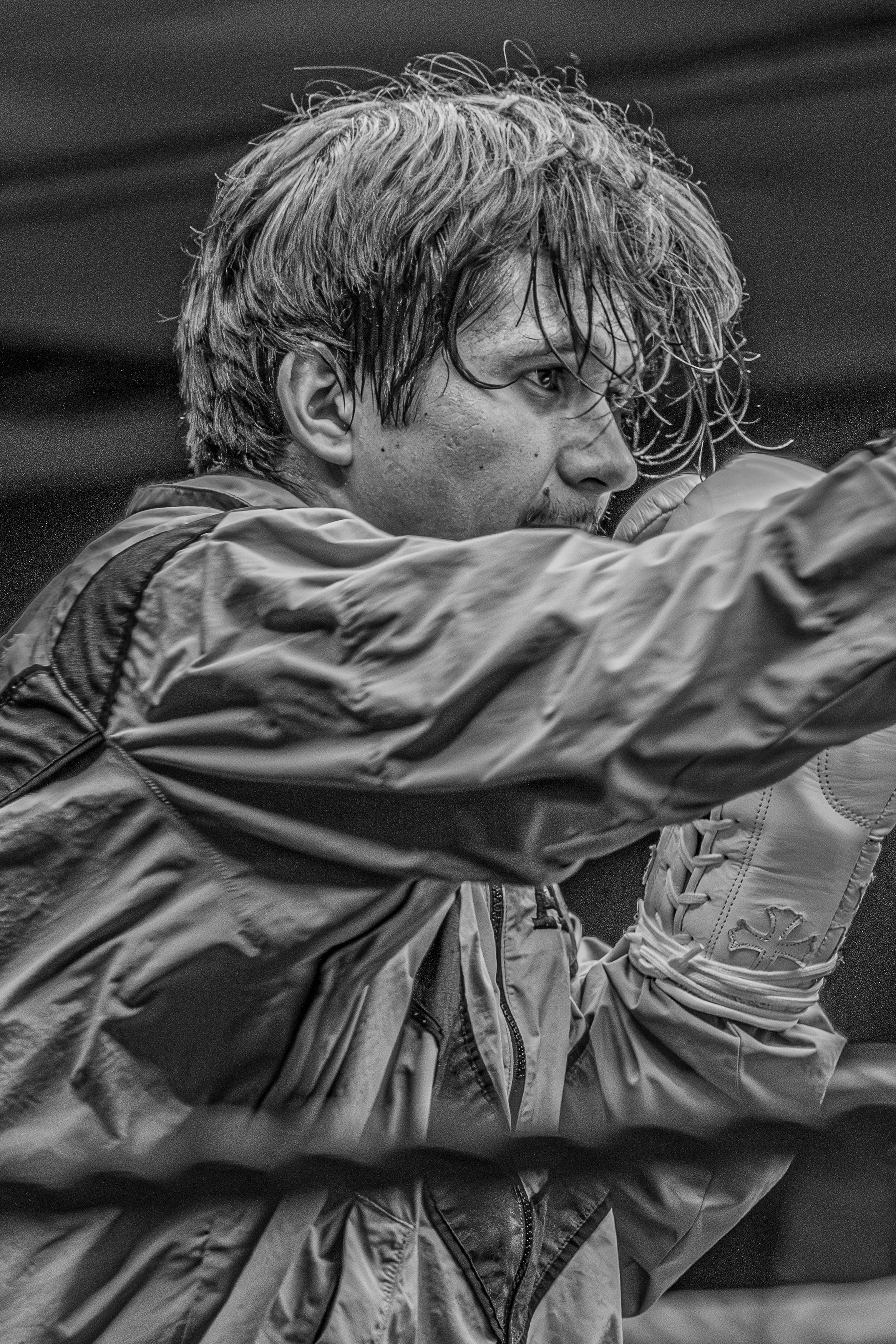 A black-and-white photo of a man with wet, disheveled hair wearing a rain jacket and a glove with a cross symbol, appearing to be training or fighting.