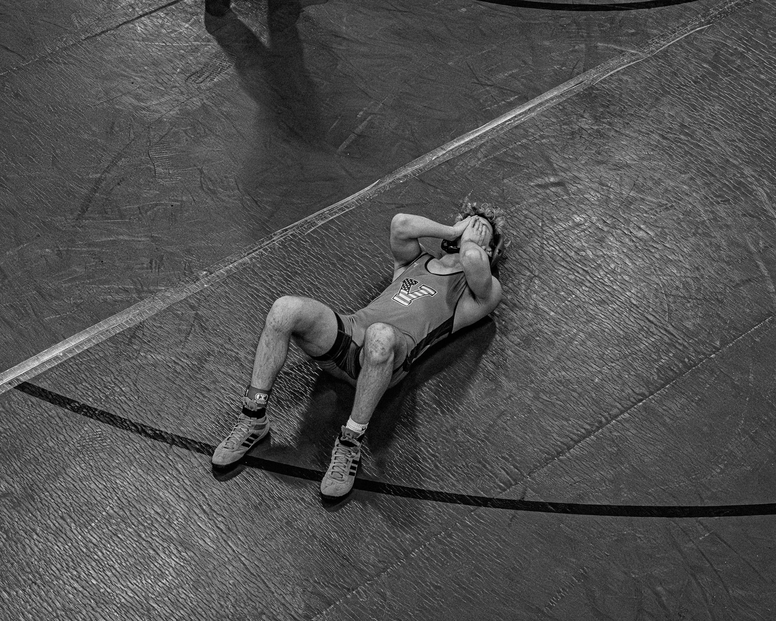 A male wrestler is lying on a wrestling mat, holding his head with both hands, showing signs of distress or exhaustion.