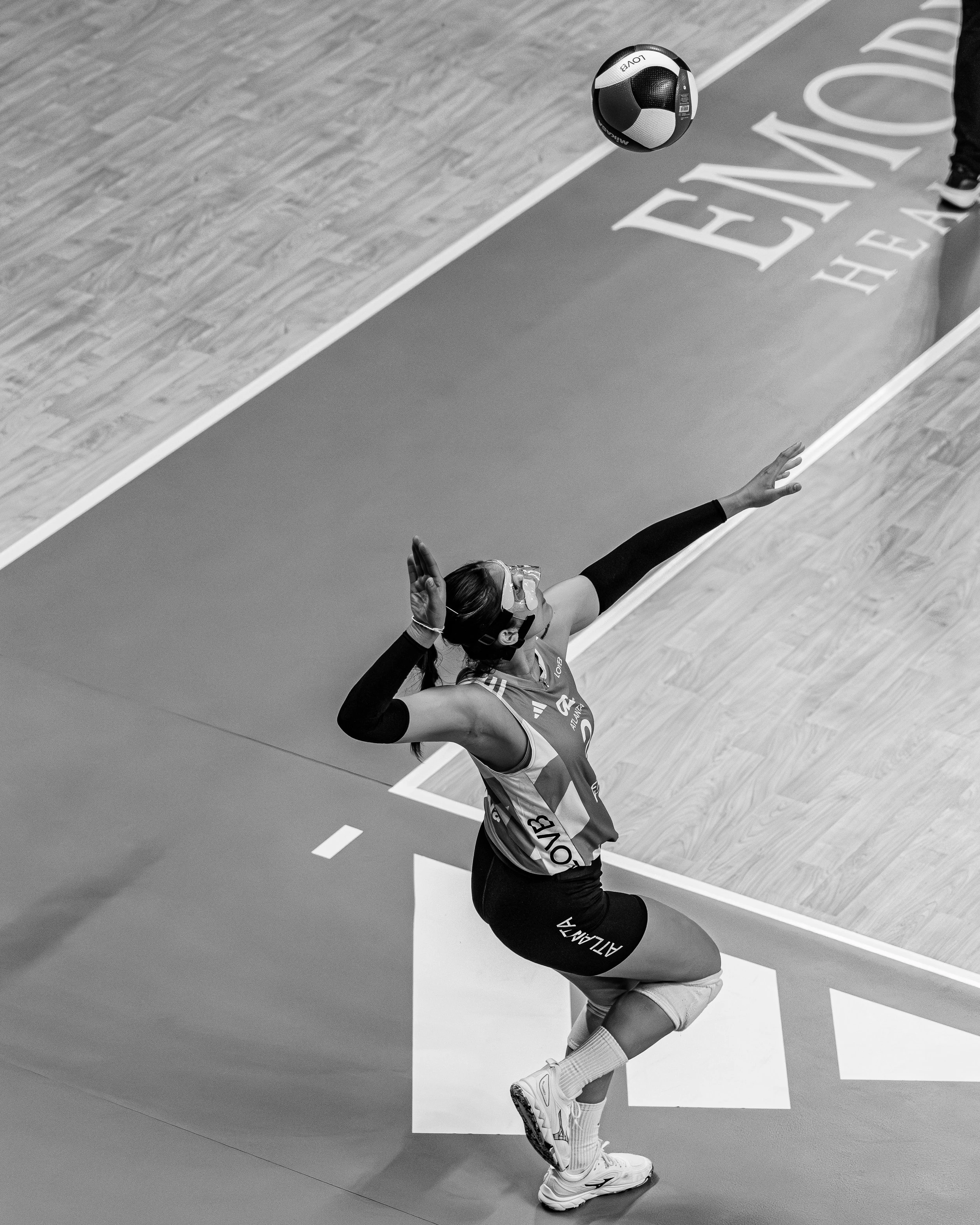A female volleyball player in a jersey, knee pads, and goggles, jumping and preparing to hit a volleyball during a game.