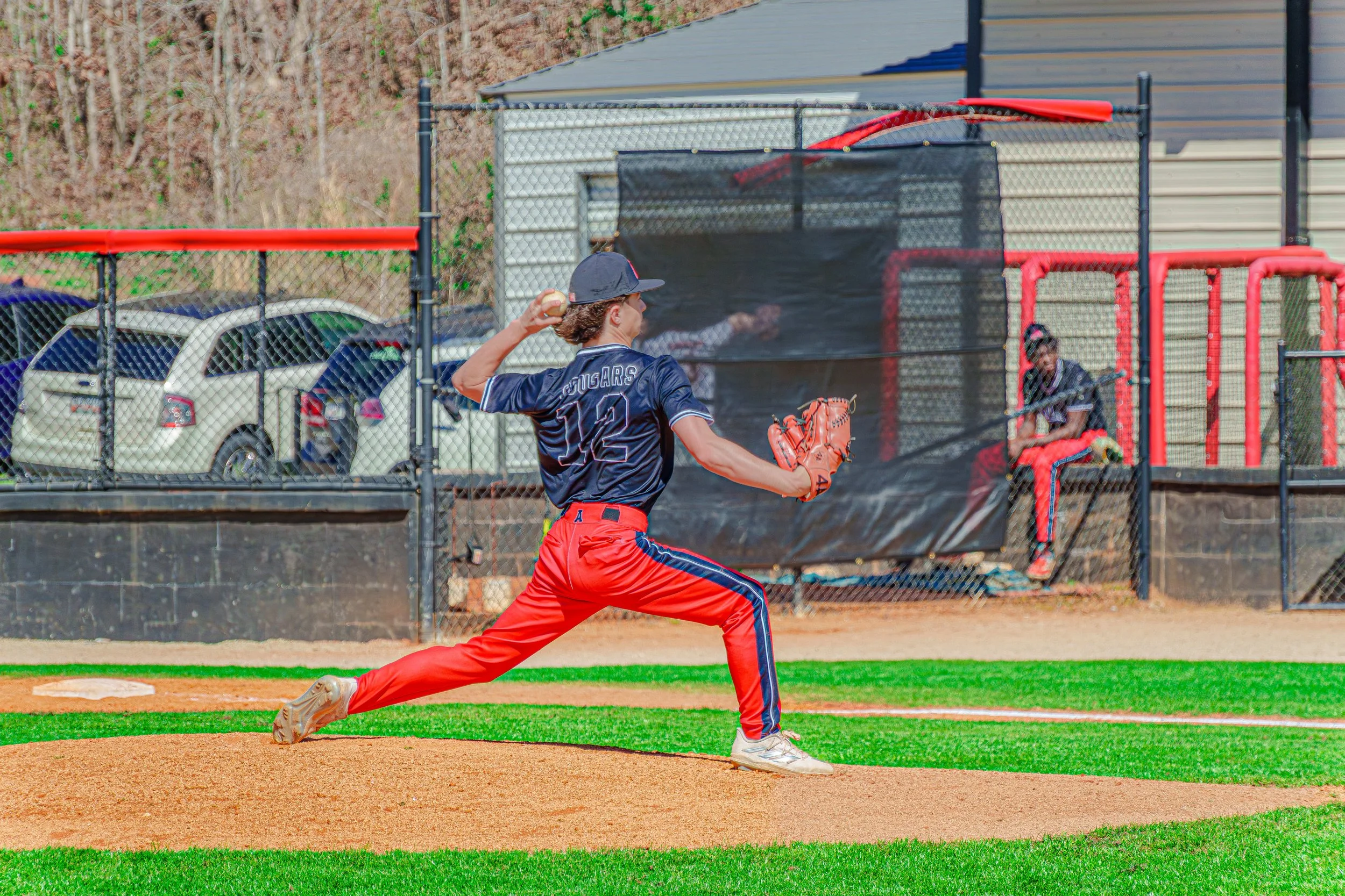 A baseball pitcher in a dark blue jersey and red pants throws a pitch on a baseball field, with a catcher sitting behind home plate.