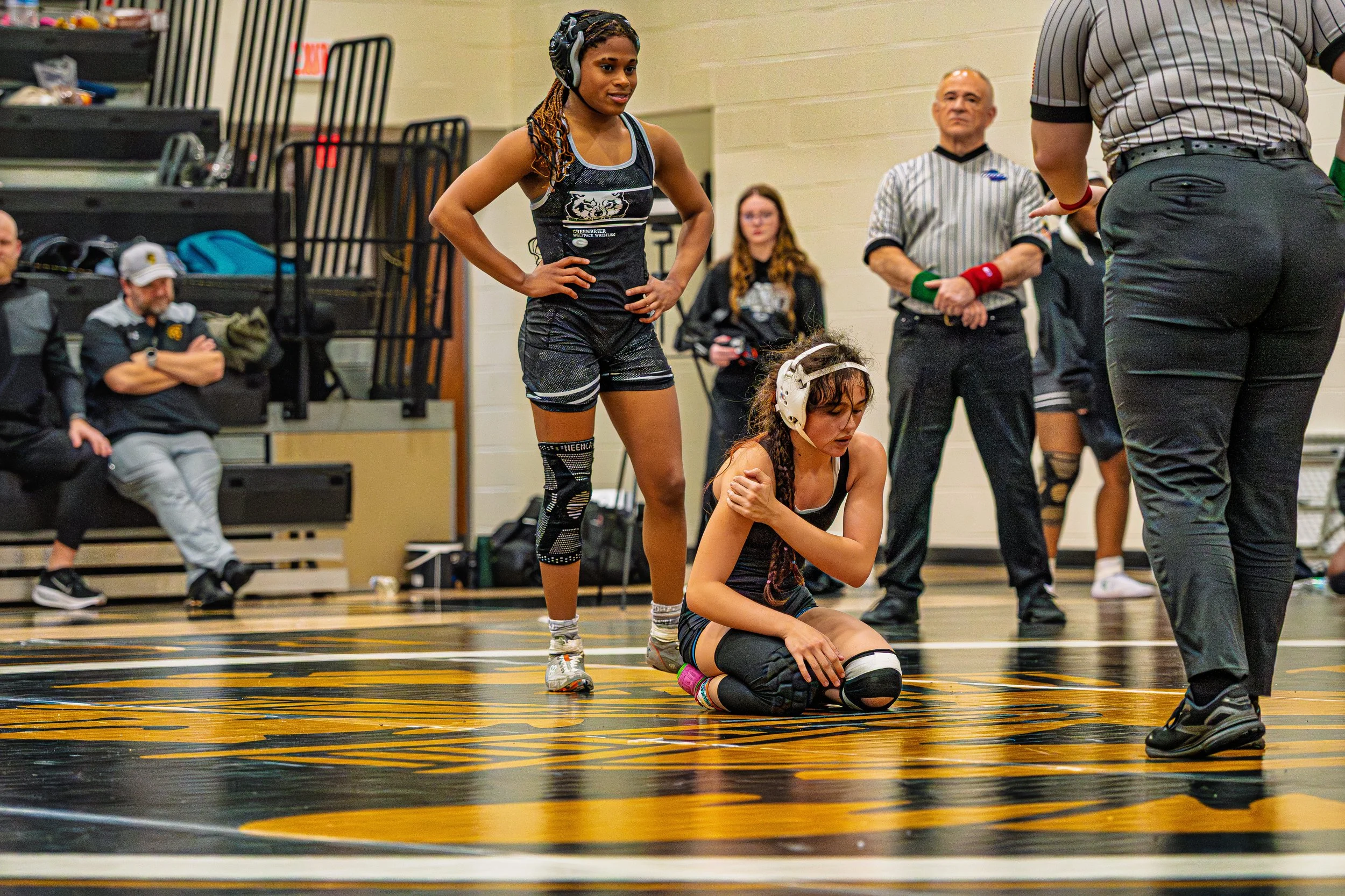 A young female wrestler kneeling on the mat, clutching her knee, with another female wrestler standing nearby and a referee in front of her during a wrestling match in a gymnasium.