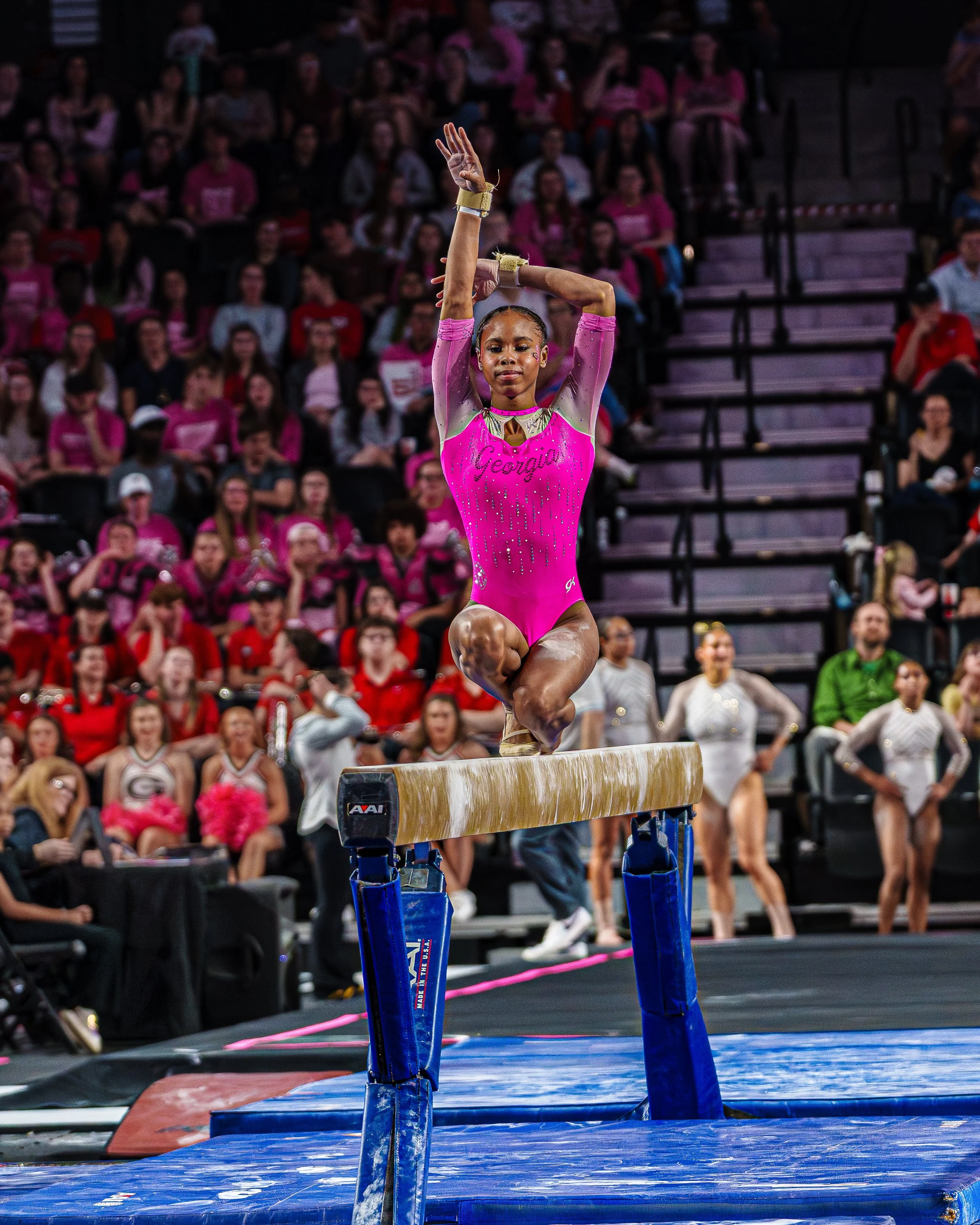 A young female gymnast in a pink leotard performing a move on the balance beam during a gymnastics competition, with an audience watching in the background.