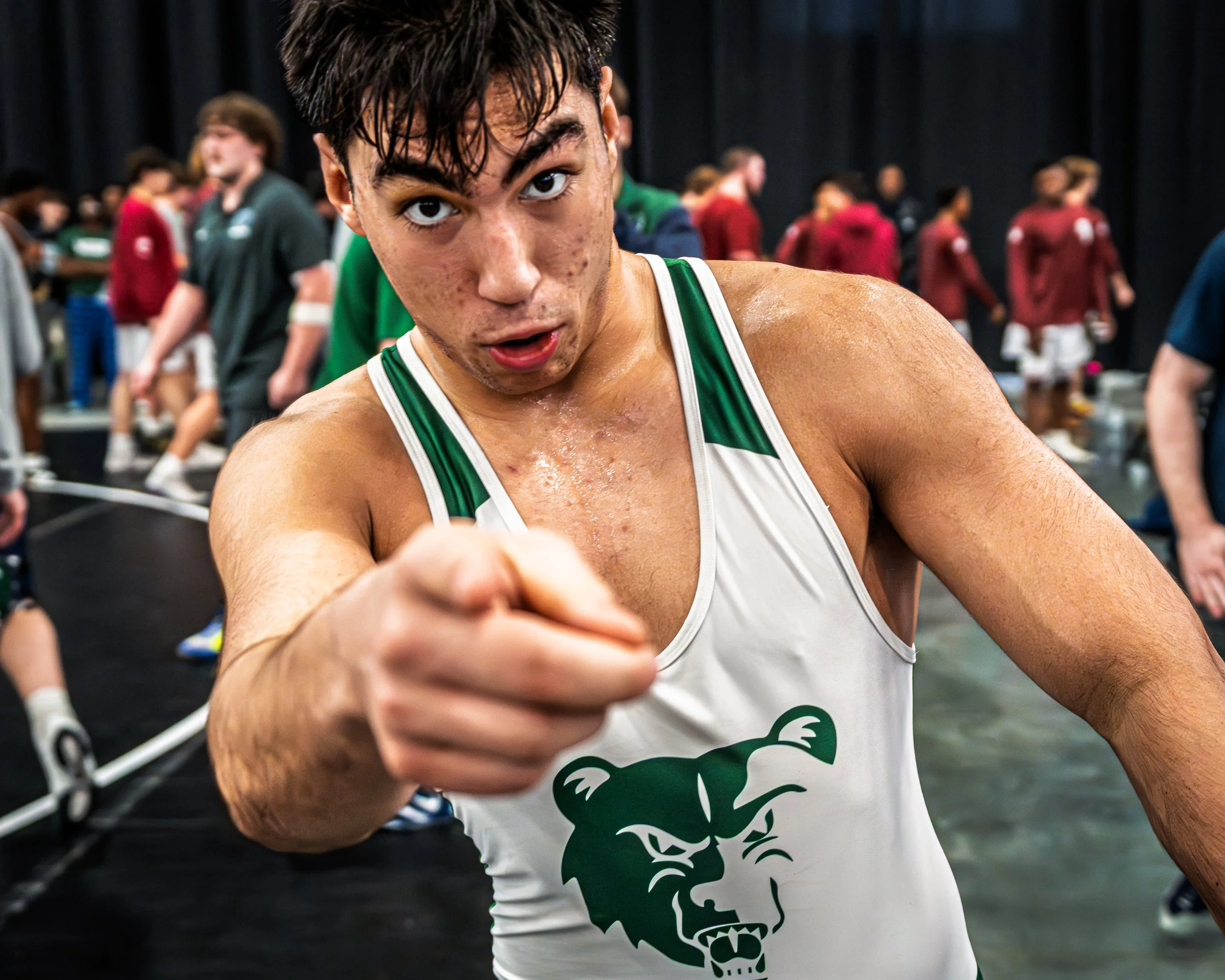 A young male athlete in a white and green track uniform pointing at the camera, sweating, with a crowd of people in the background.