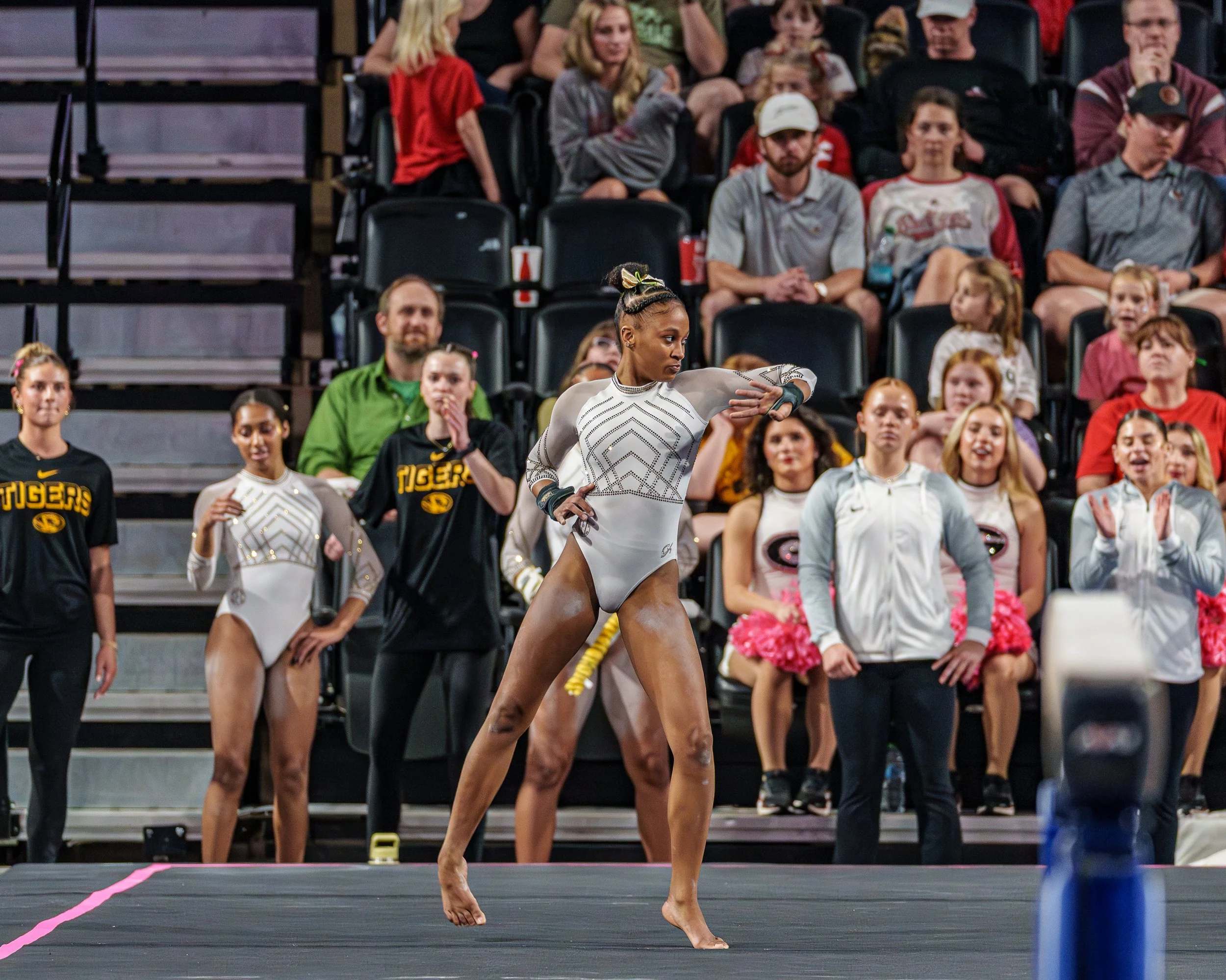 A female gymnast in a white leotard performs on the floor in front of an audience, with other gymnasts and spectators watching.