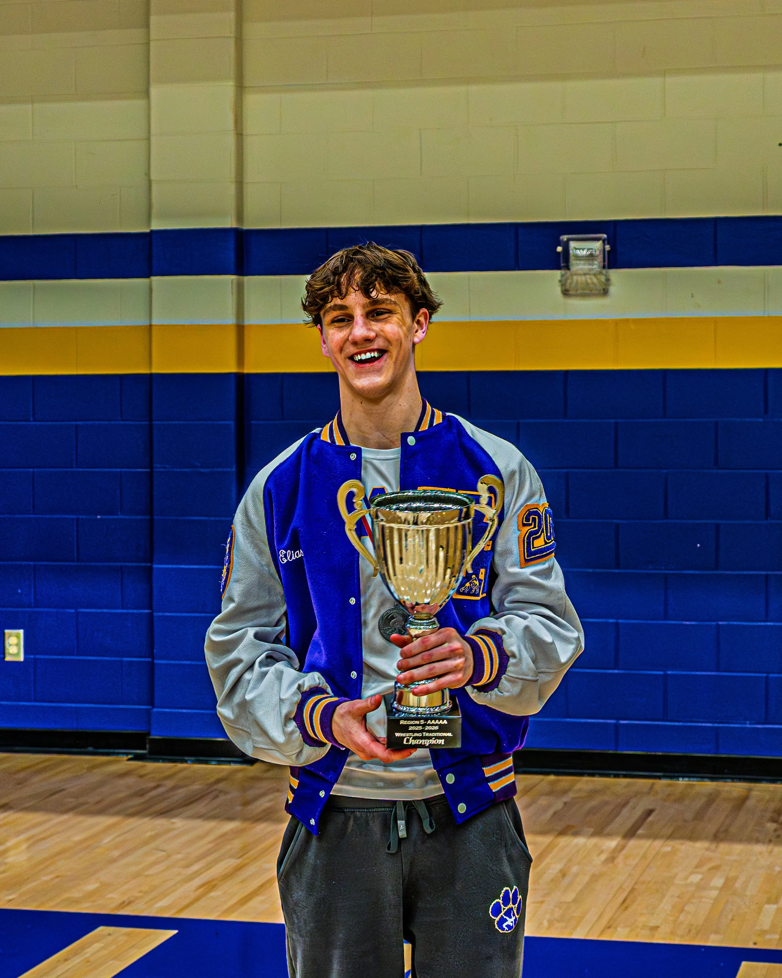 Teenager smiling and holding a trophy in a gymnasium, wearing a varsity jacket and sweatpants with a paw print logo.