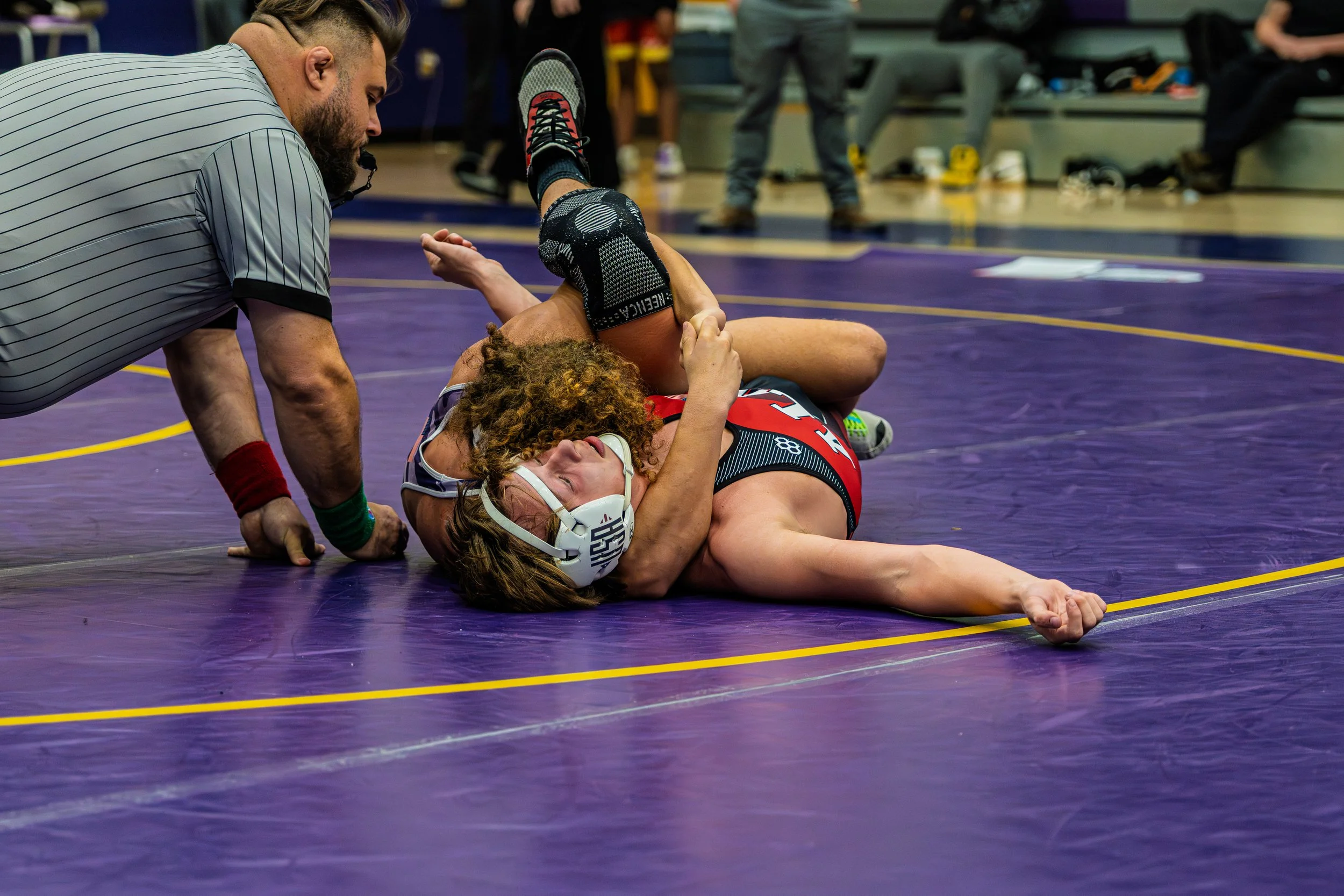 A wrestling match with a female wrestler in a red and black singlet on the mat, being pinning by her opponent, while the referee crouches beside them.