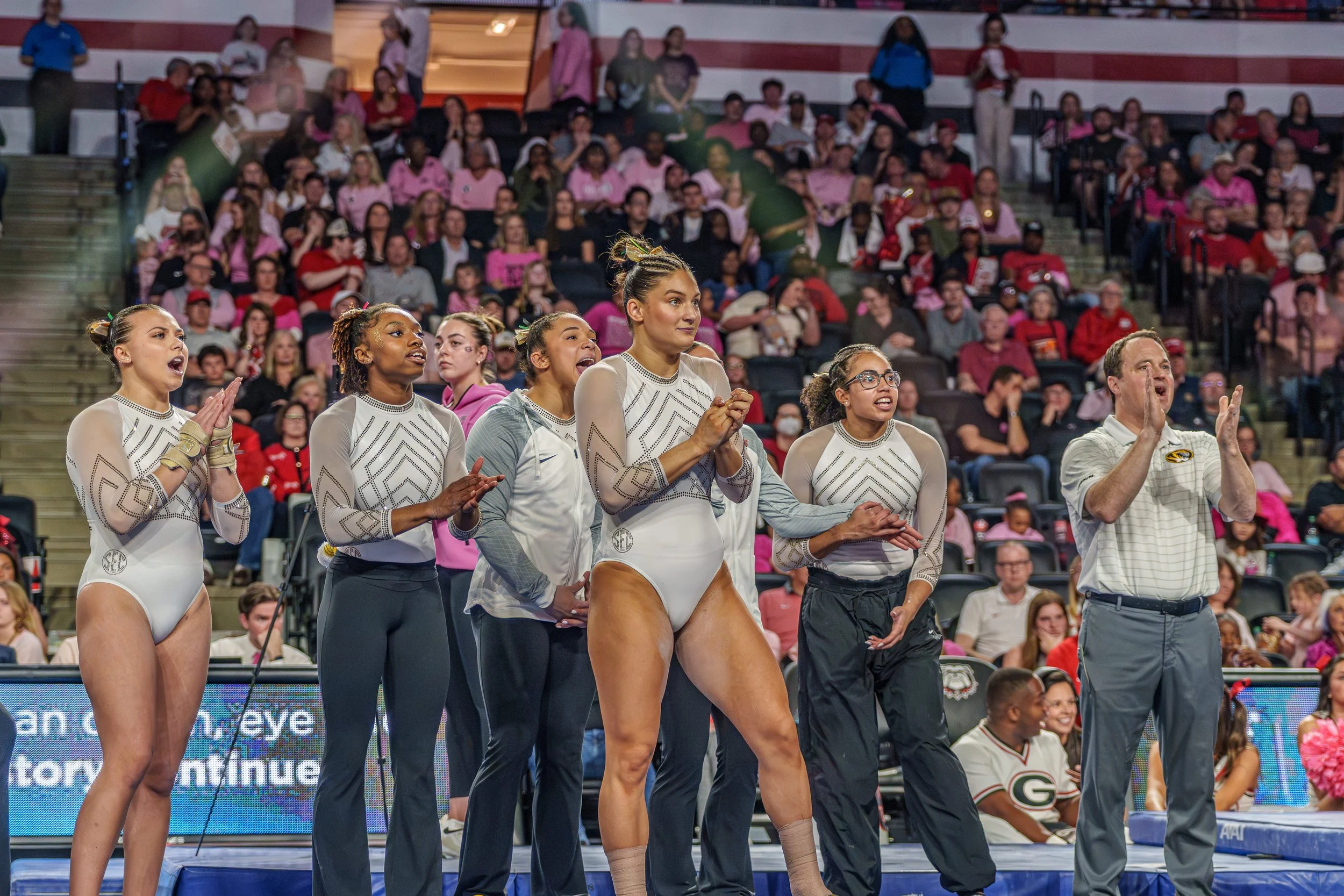 A woman gymnast in a white leotard with gray and black accents standing on a gymnastics mat, surrounded by four women and one man, all clapping and cheering. The audience in the background is seated in an arena.