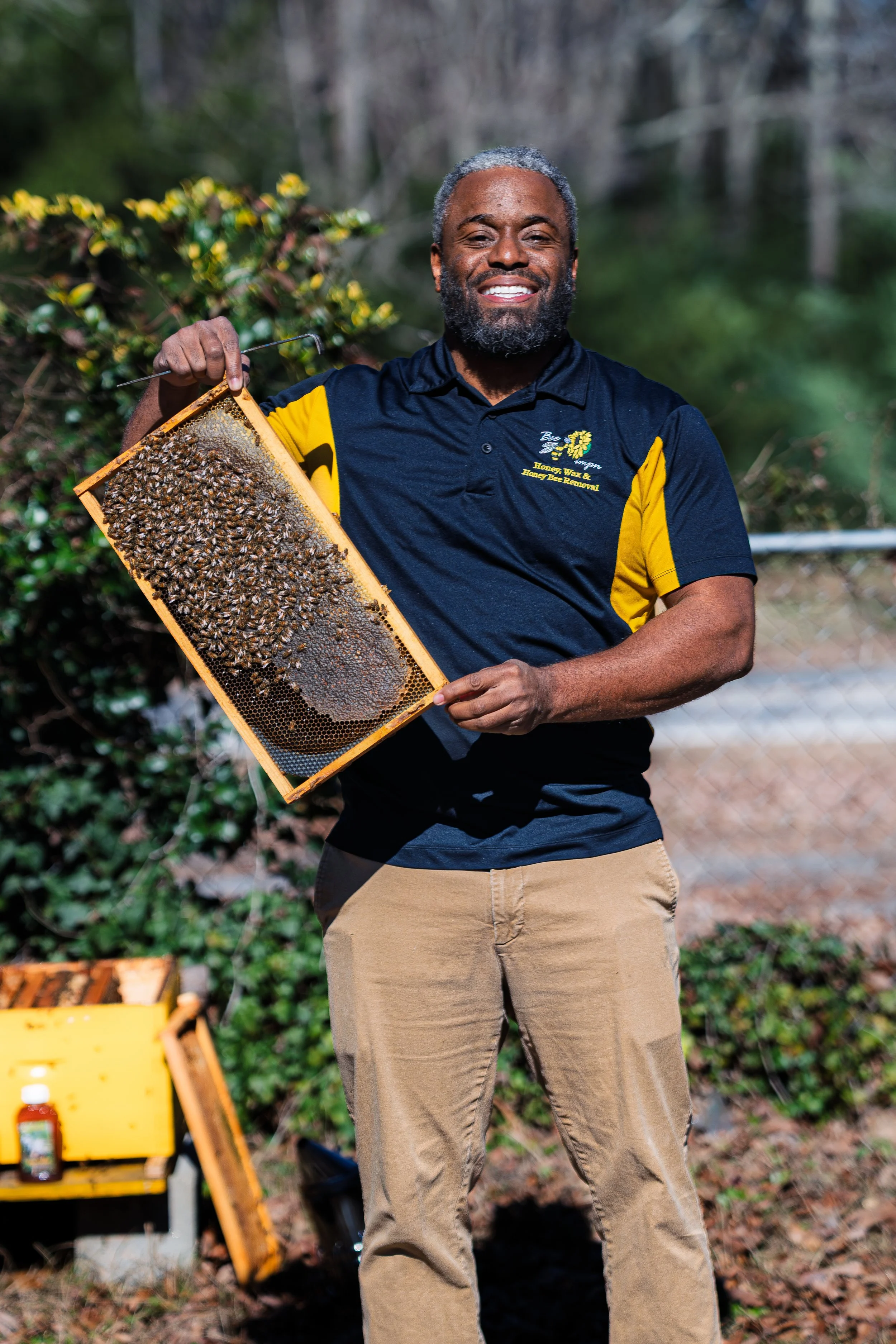A man smiling outdoors while holding a honeycomb frame filled with bees and honey.