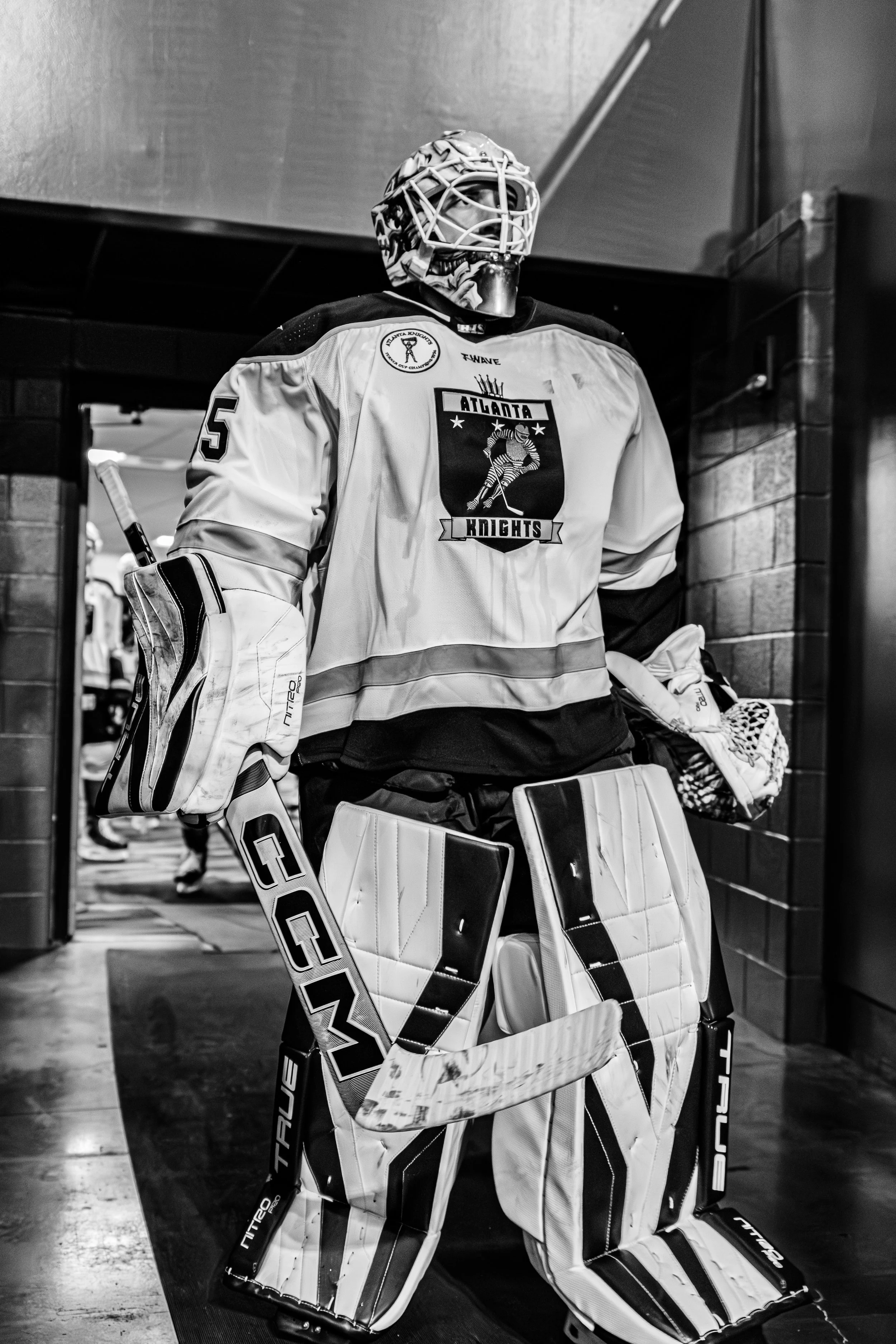 Black and white photo of a hockey goalie in full gear with a jersey labeled 'Atlanta Knights' standing in a locker room.