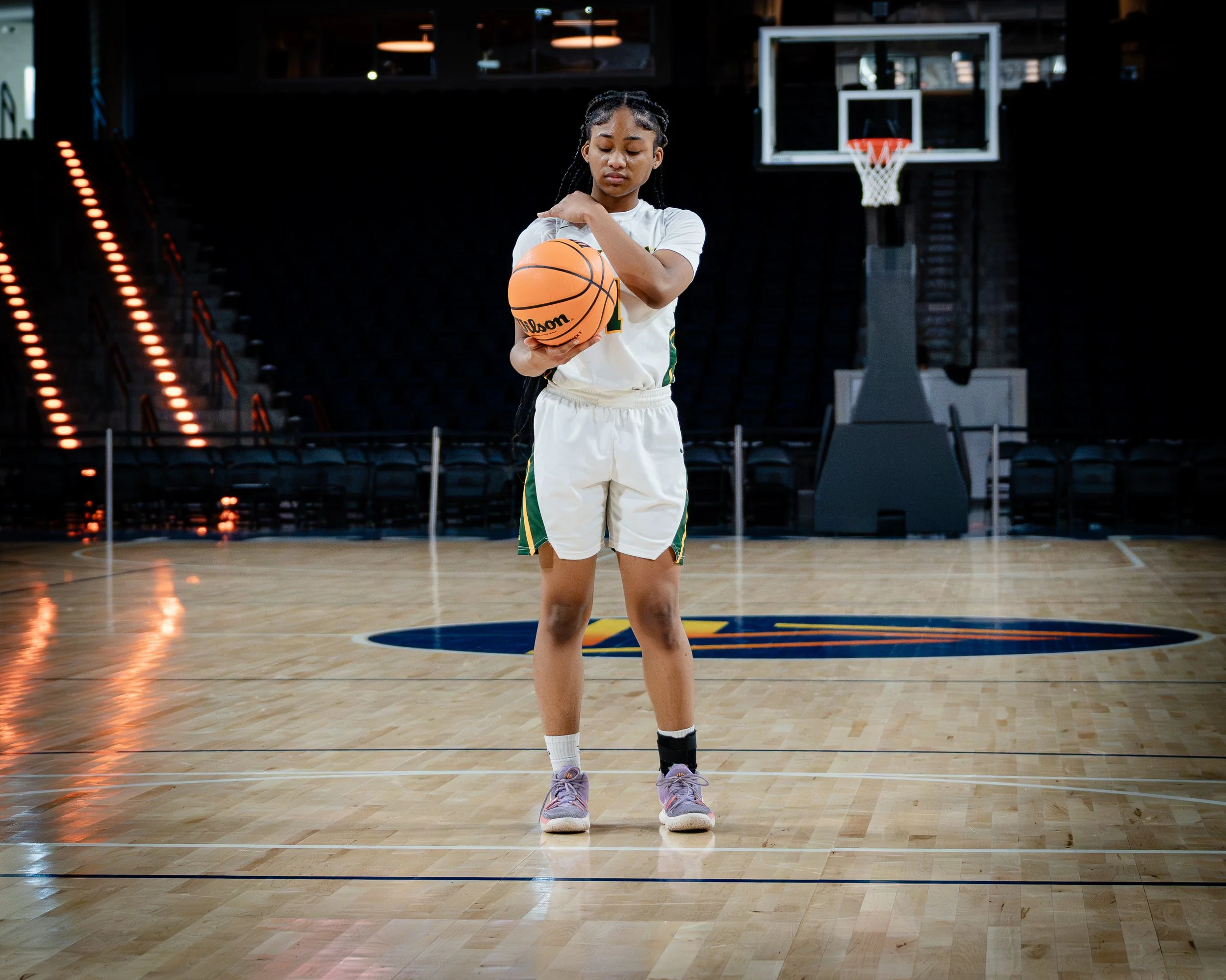 Young female basketball player standing on a basketball court holding a basketball, with a basketball hoop in the background.