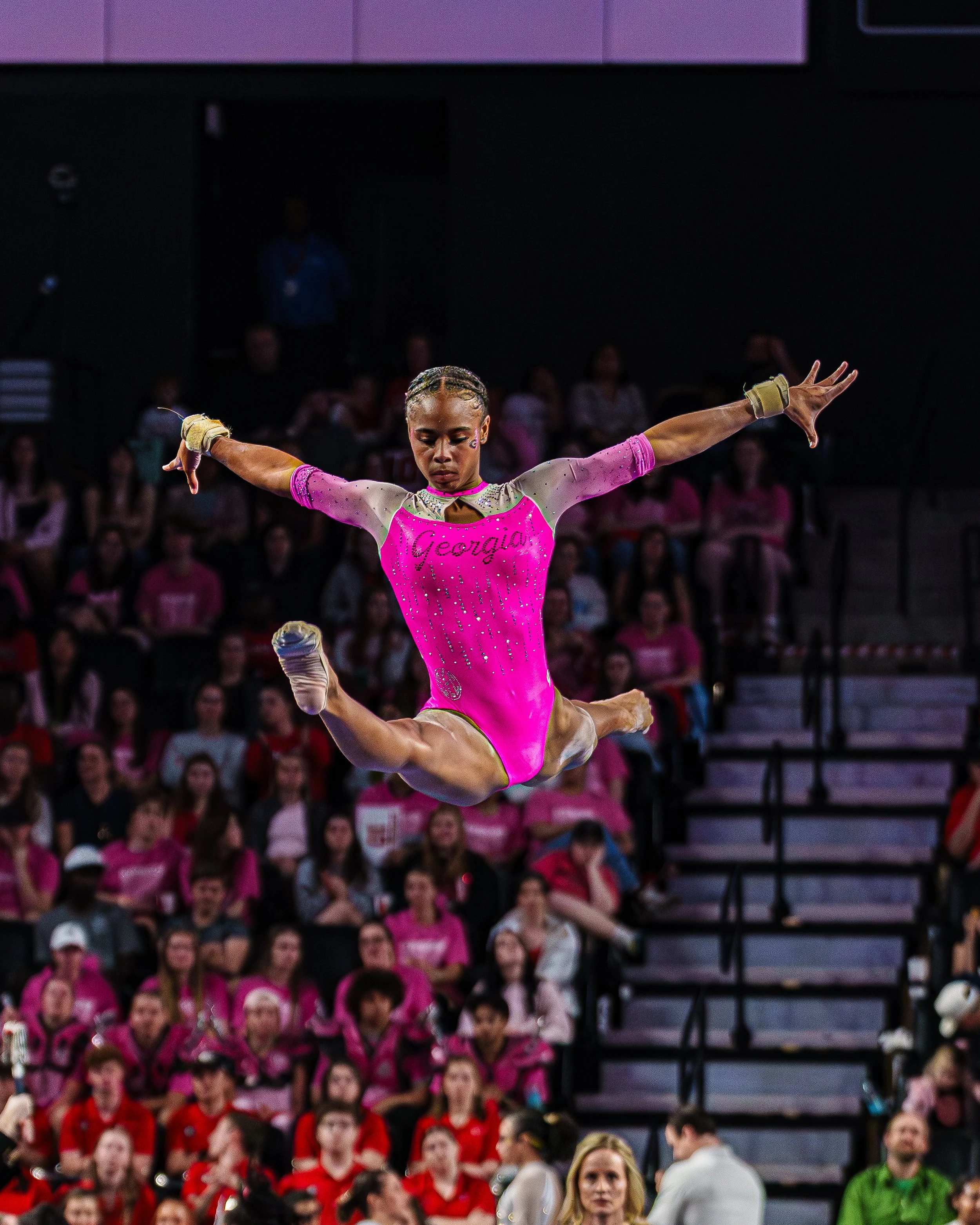 A female gymnast in pink leotard performing a leap during a gymnastics competition with a crowd in the background.