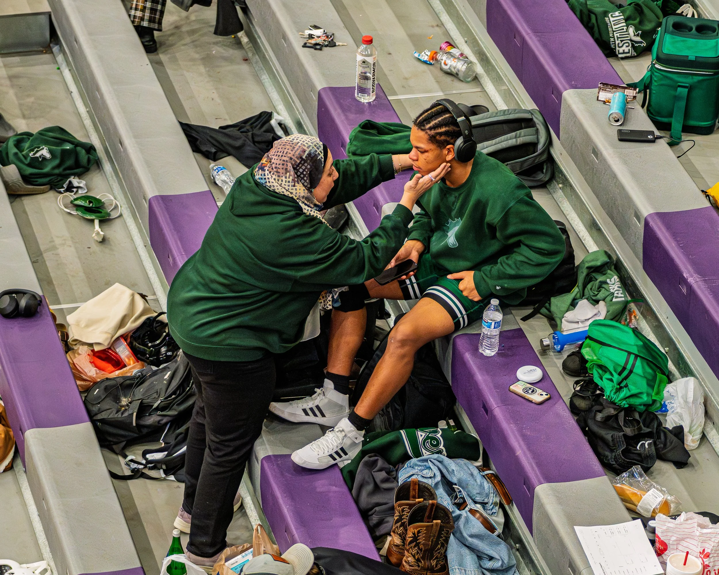 A woman in a hijab tending to a young male athlete with cornrows wearing headphones at a sports venue, likely a basketball game.