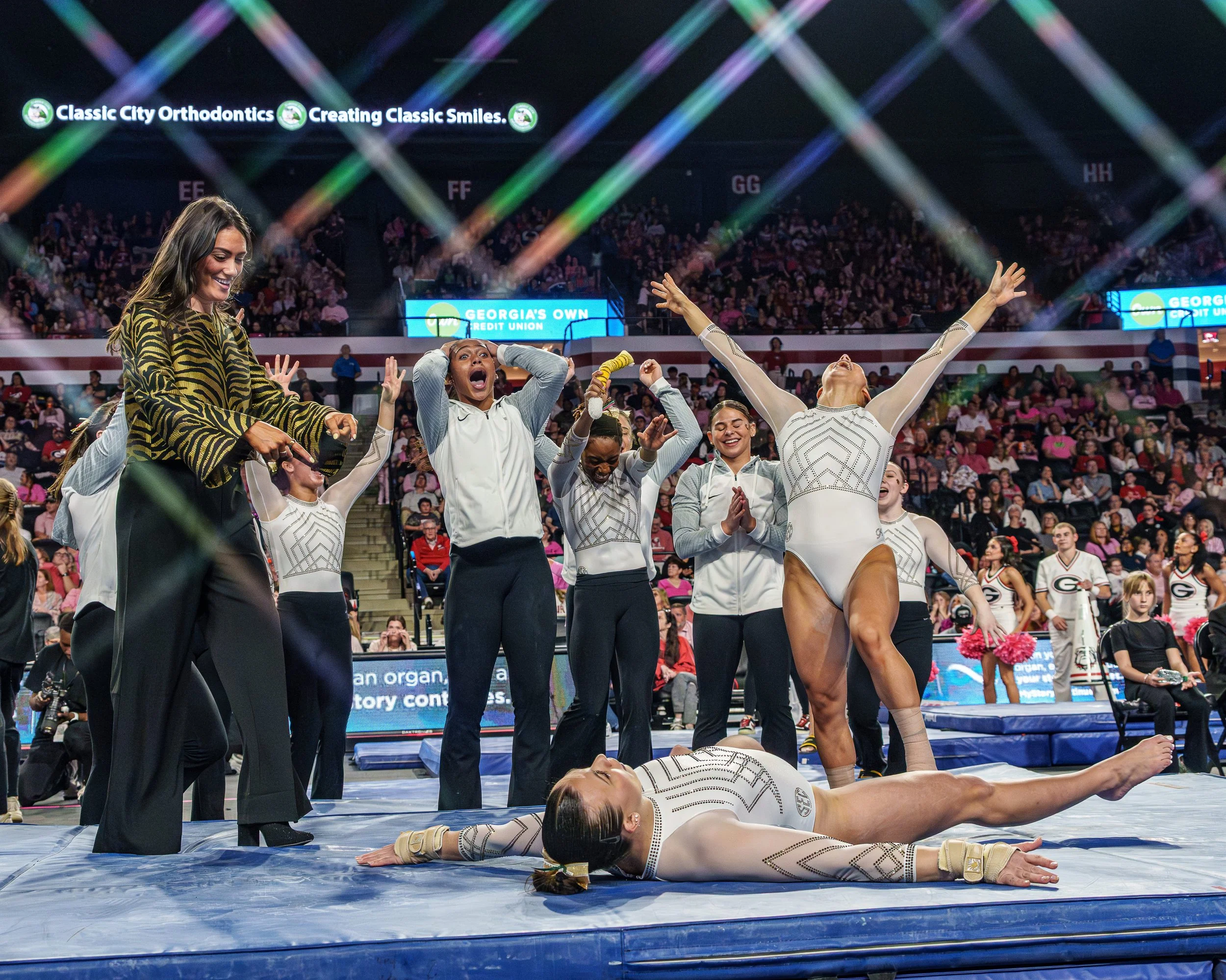 Female gymnasts celebrating after a successful routine on the balance beam during a gymnastics competition, with coaches and spectators present.