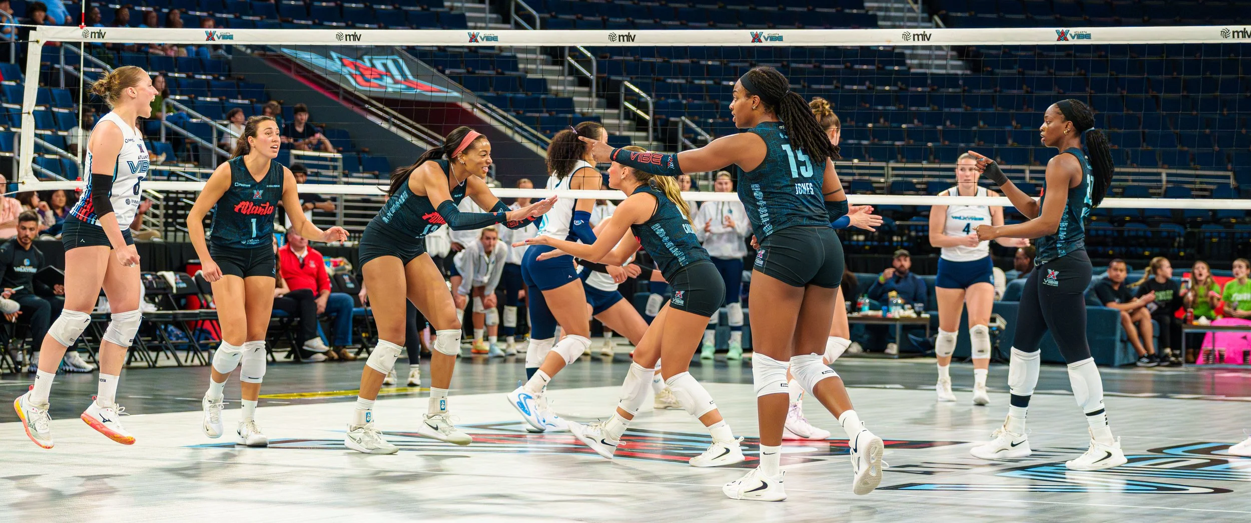 Women's volleyball team celebrating during a match, with players wearing black and white uniforms on an indoor court.