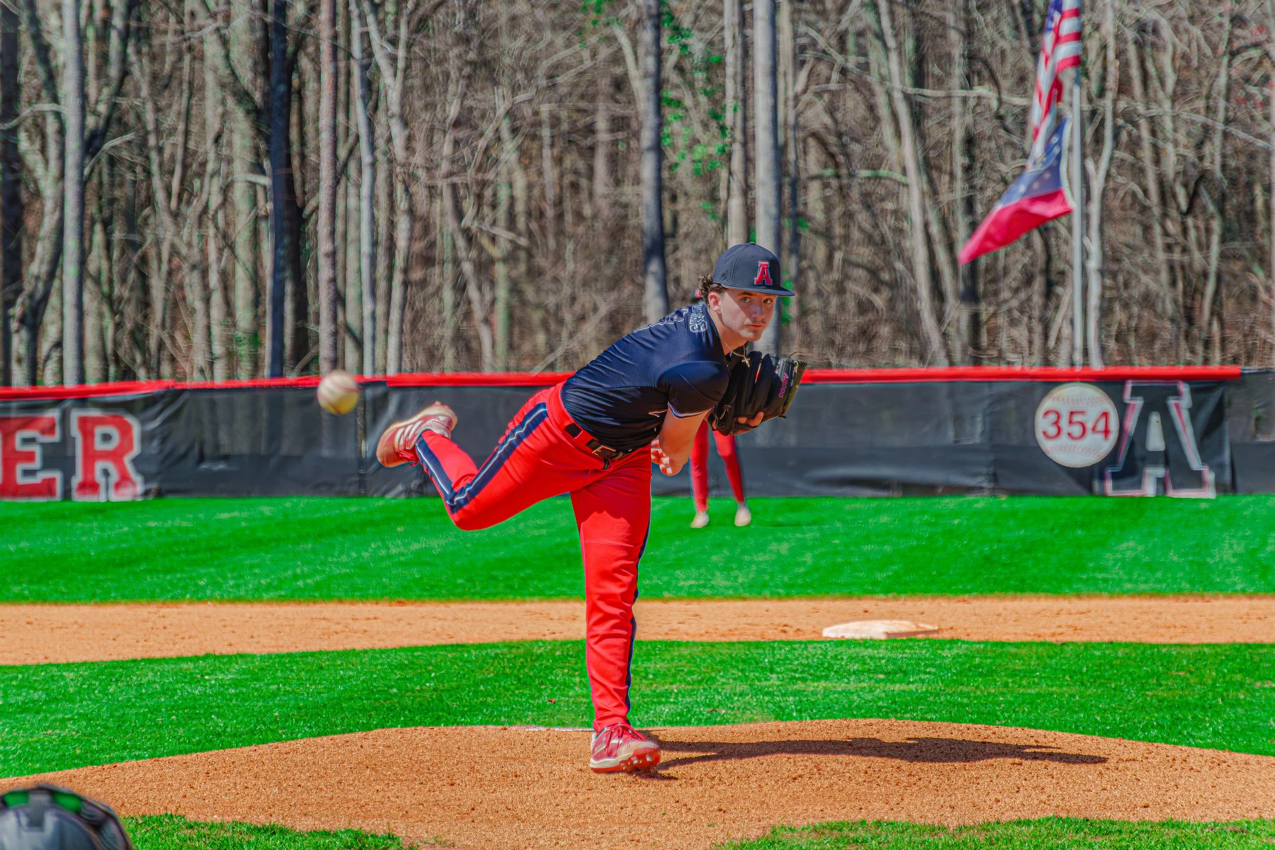 A young baseball pitcher in red and navy uniform mid-throw on a baseball field.