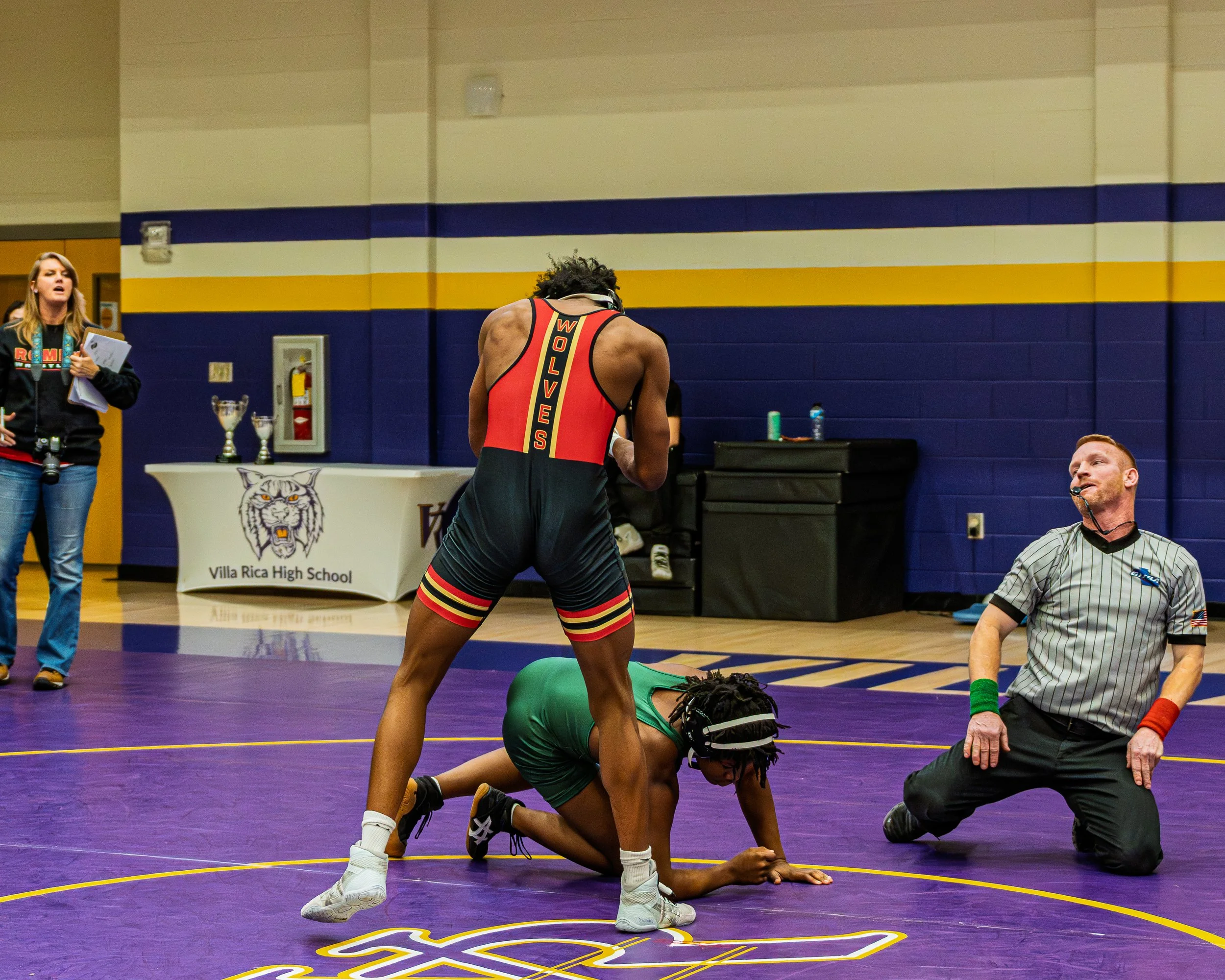 Two high school wrestlers in a match on a purple mat, one in a red and black uniform labeled 'Wolves' standing over another in a green uniform who is on all fours. A referee kneels beside them, observing. In the background, a table with trophies and 