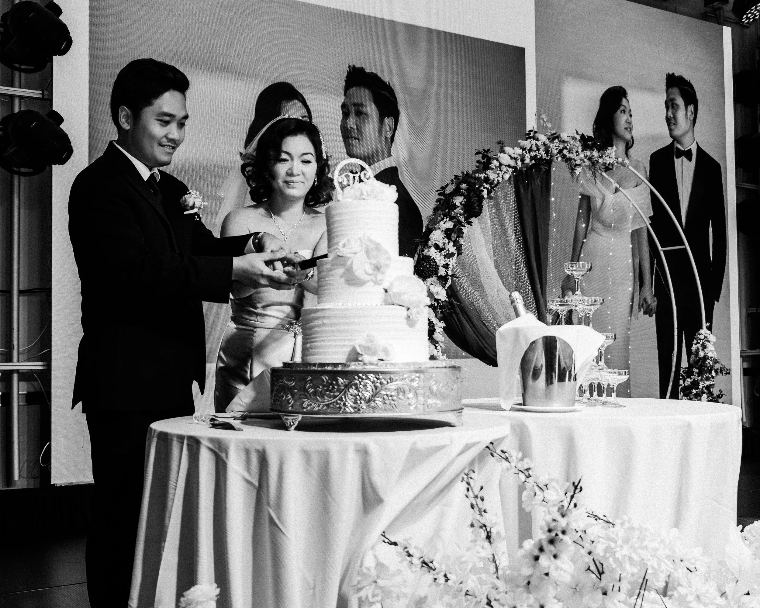 Black and white photo of a wedding celebration with a couple cutting a tiered wedding cake. Three other people are standing behind them, two women and one man, on a decorated stage with a large backdrop. The table in front of them has a bucket, a cak