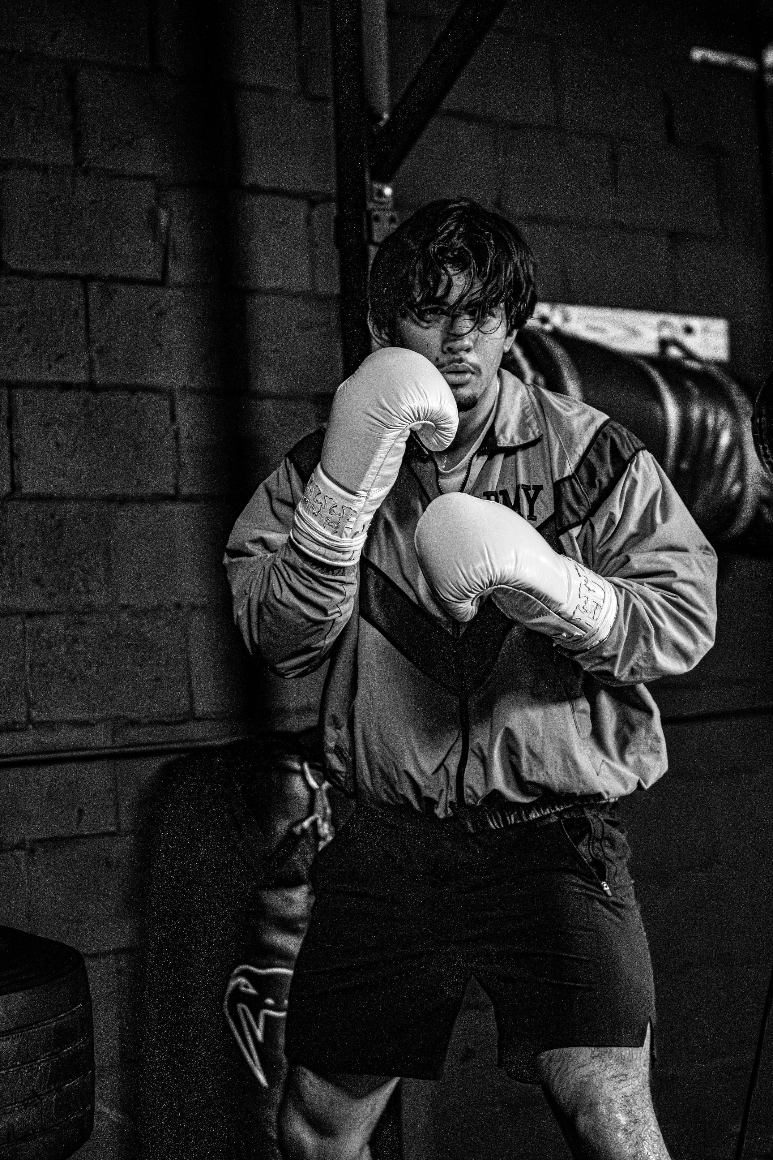A young man with dark hair and glasses wearing boxing gloves and athletic clothing, posing in a boxing gym against a brick wall.
