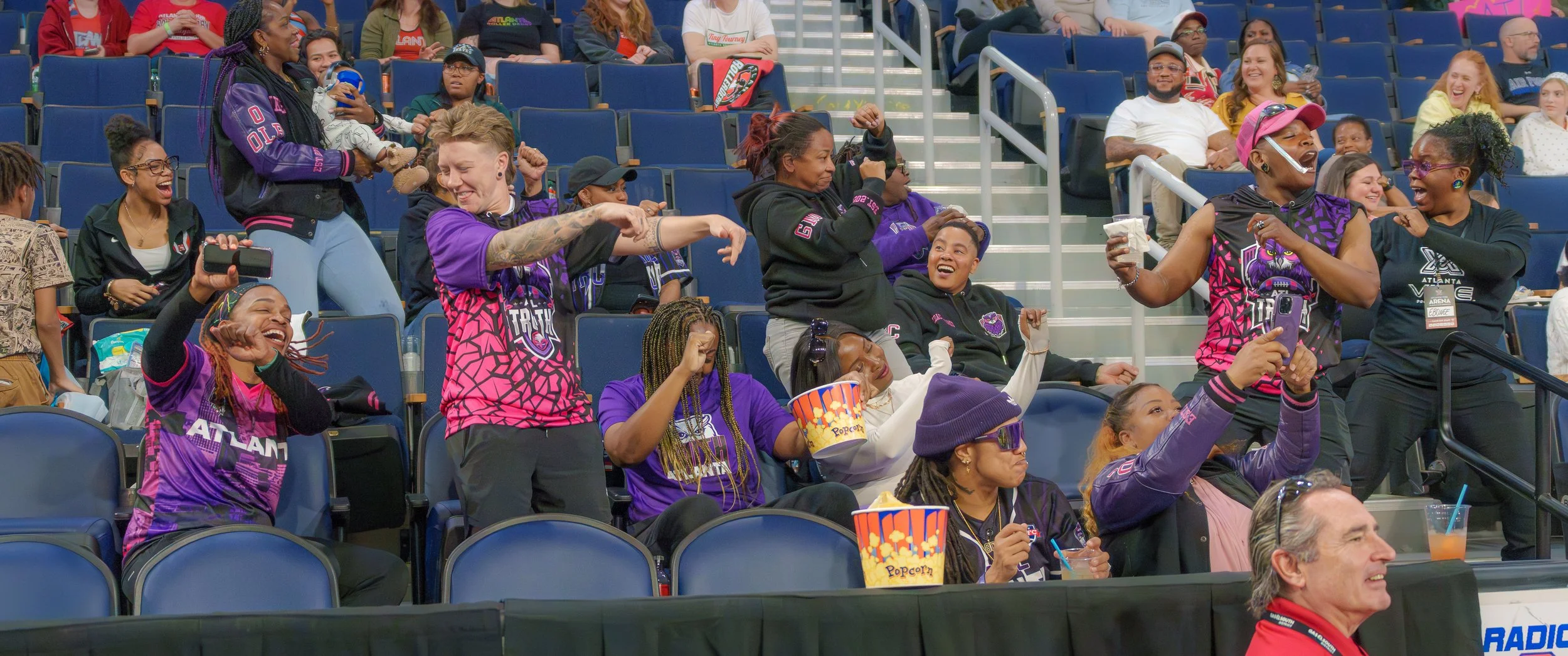 Group of excited women in Atlanta Falcons team apparel at a sports event, cheering and taking photos in stadium seats.
