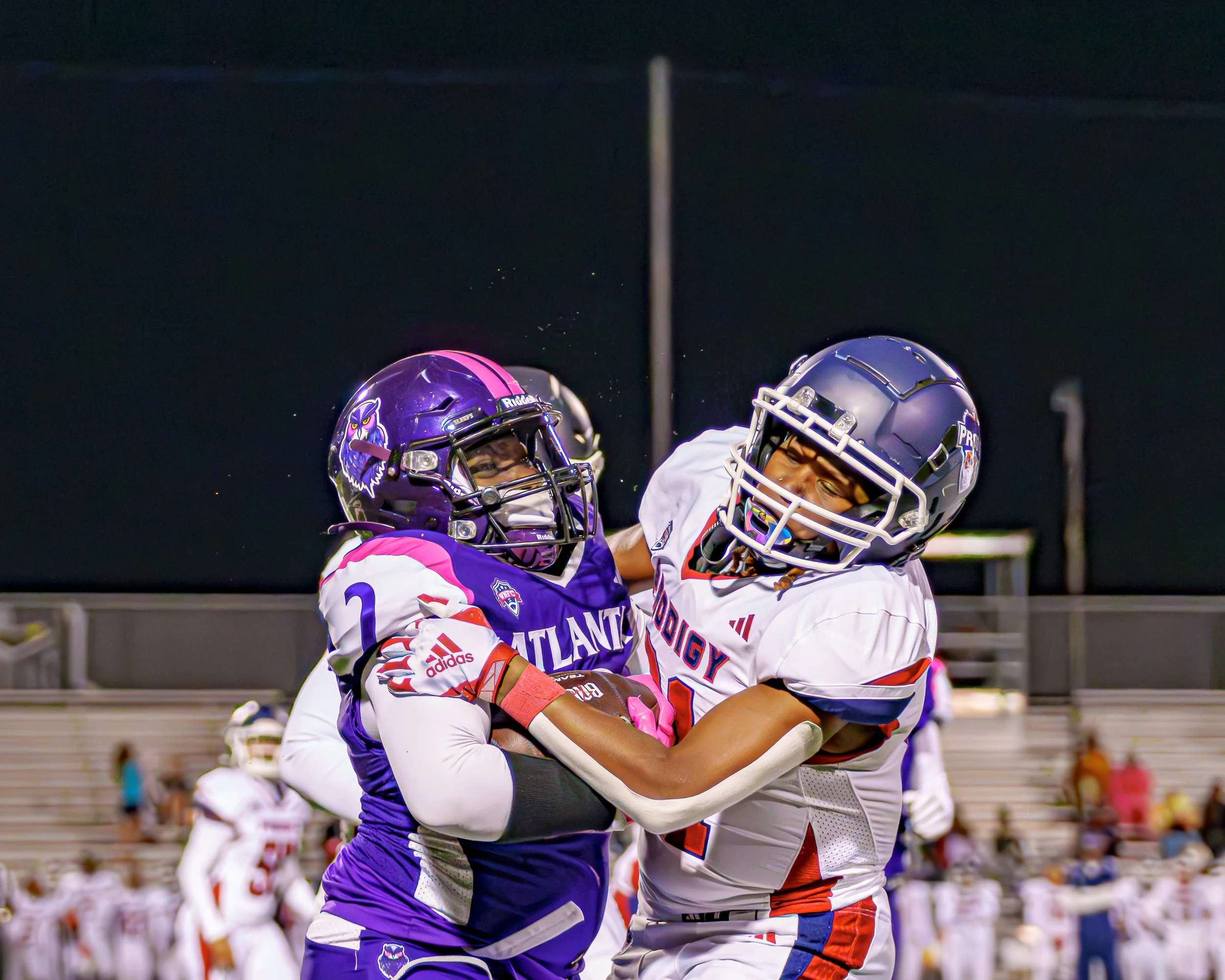 Two football players in helmets and uniforms engage in a tackle during a game at night, with fans in the stands in the background.