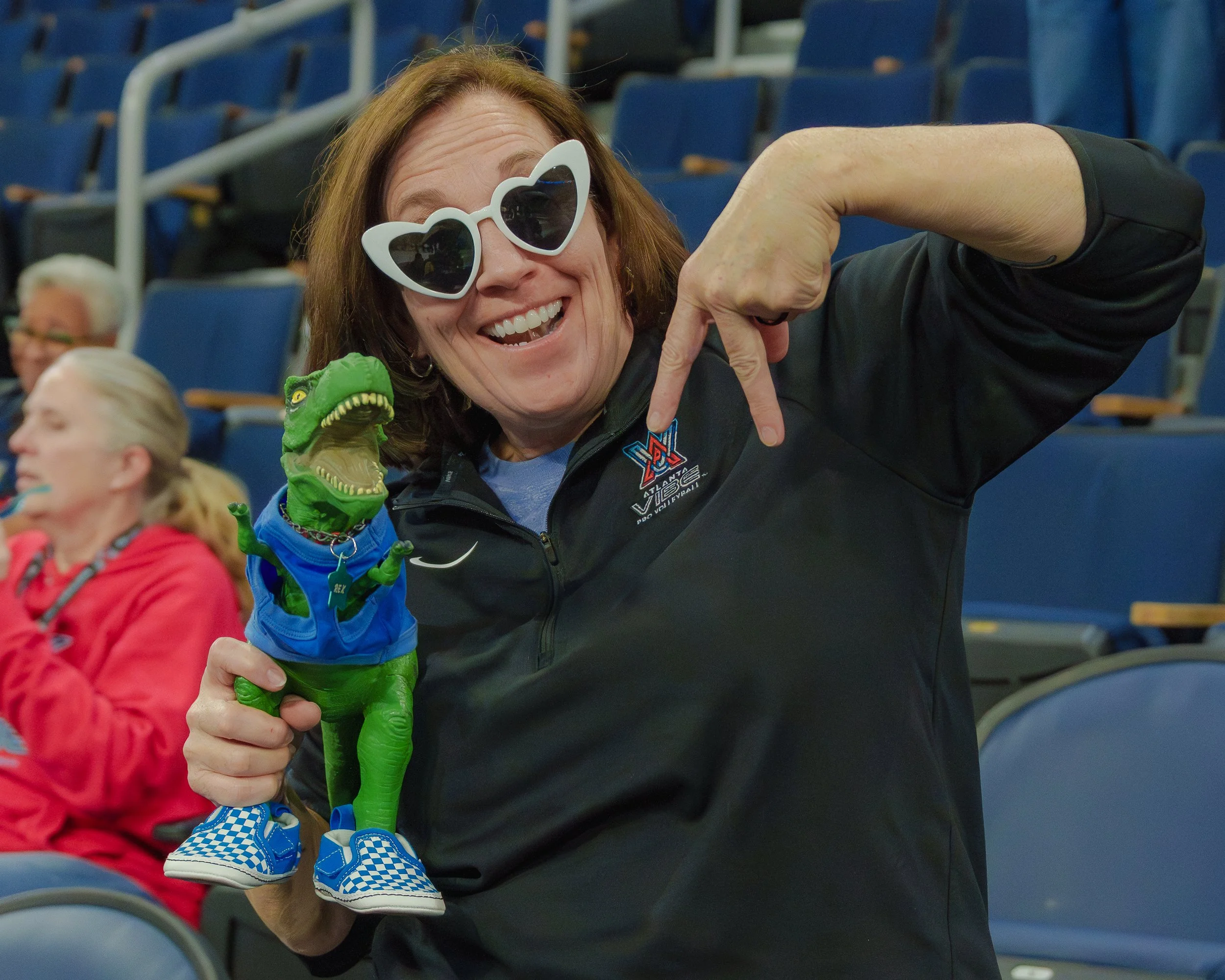 A woman wearing sunglasses and a jacket with a Volleyball logo, smiling and pointing to a dinosaur plush toy dressed in sneakers, in an indoor arena with empty blue seats.