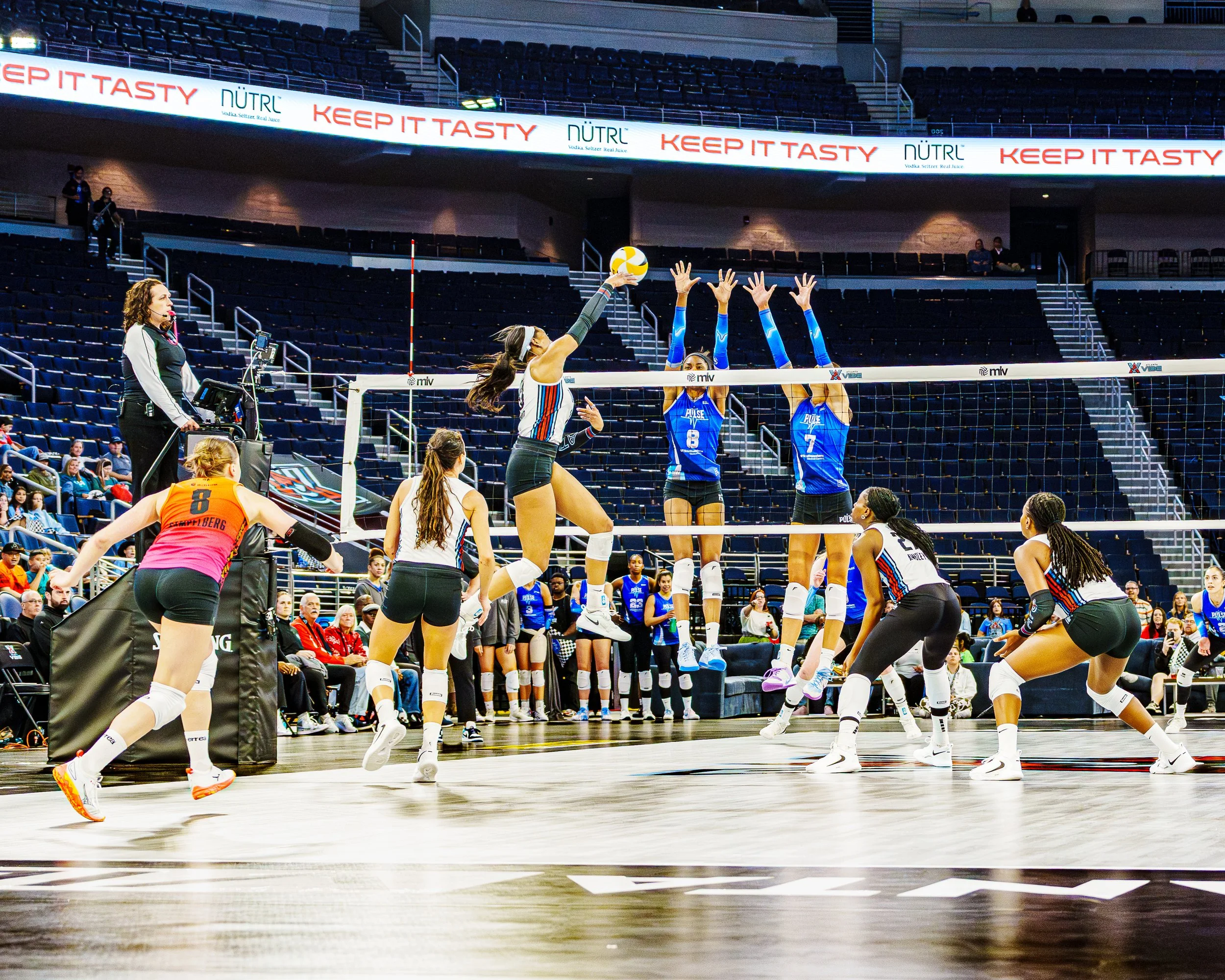 Women's volleyball game in an indoor arena with players in action. A player from one team is jumping to strike the ball, while three players from the opposing team are jumping to block. Spectators are visible in the background.