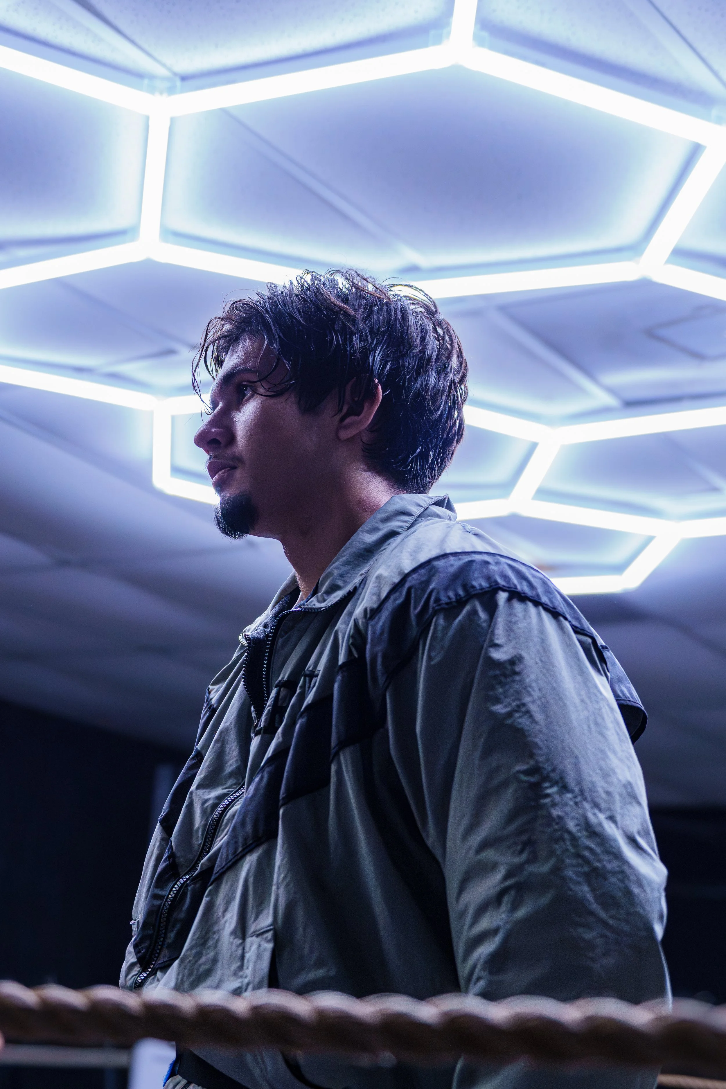 A man with wet hair and a goatee, wearing a gray jacket, standing indoors under a futuristic ceiling light installation with geometric shapes, illuminated with bright white light.