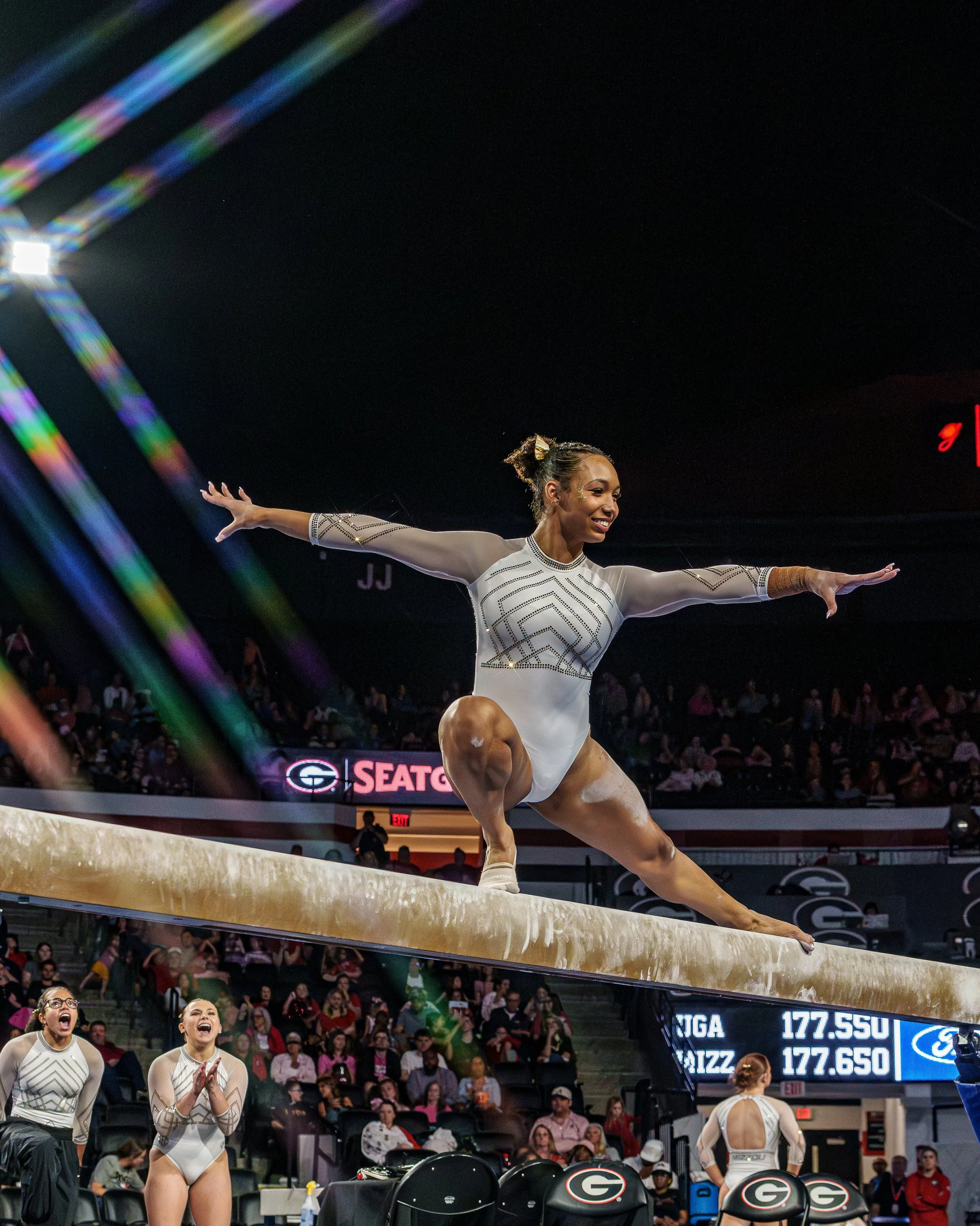 A female gymnast in a white leotard performing a split on the balance beam during a gymnastics competition, with spectators in the background and judges watching.