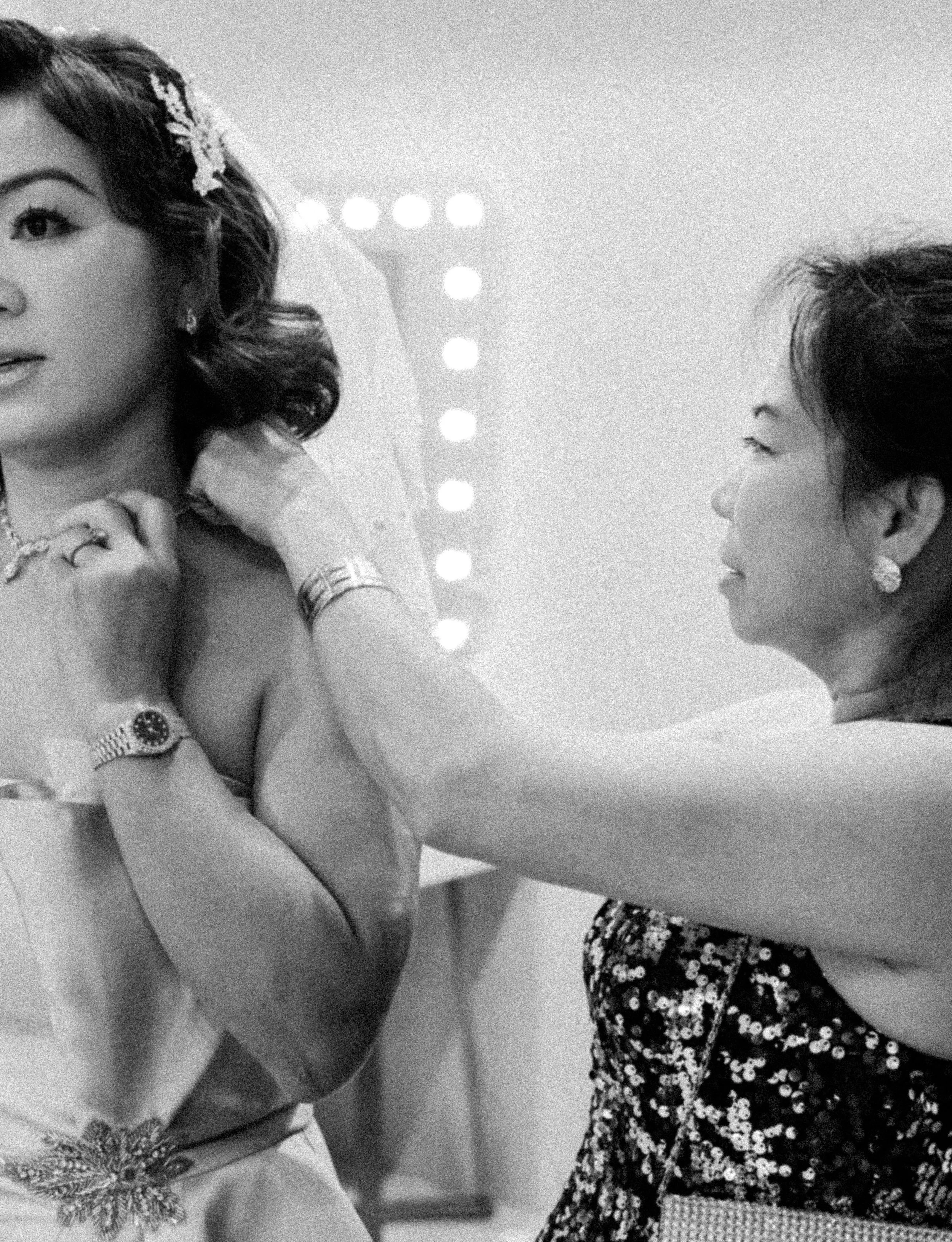 A woman is getting ready, with another woman helping her adjust her dress, in a dressing room with mirror lights