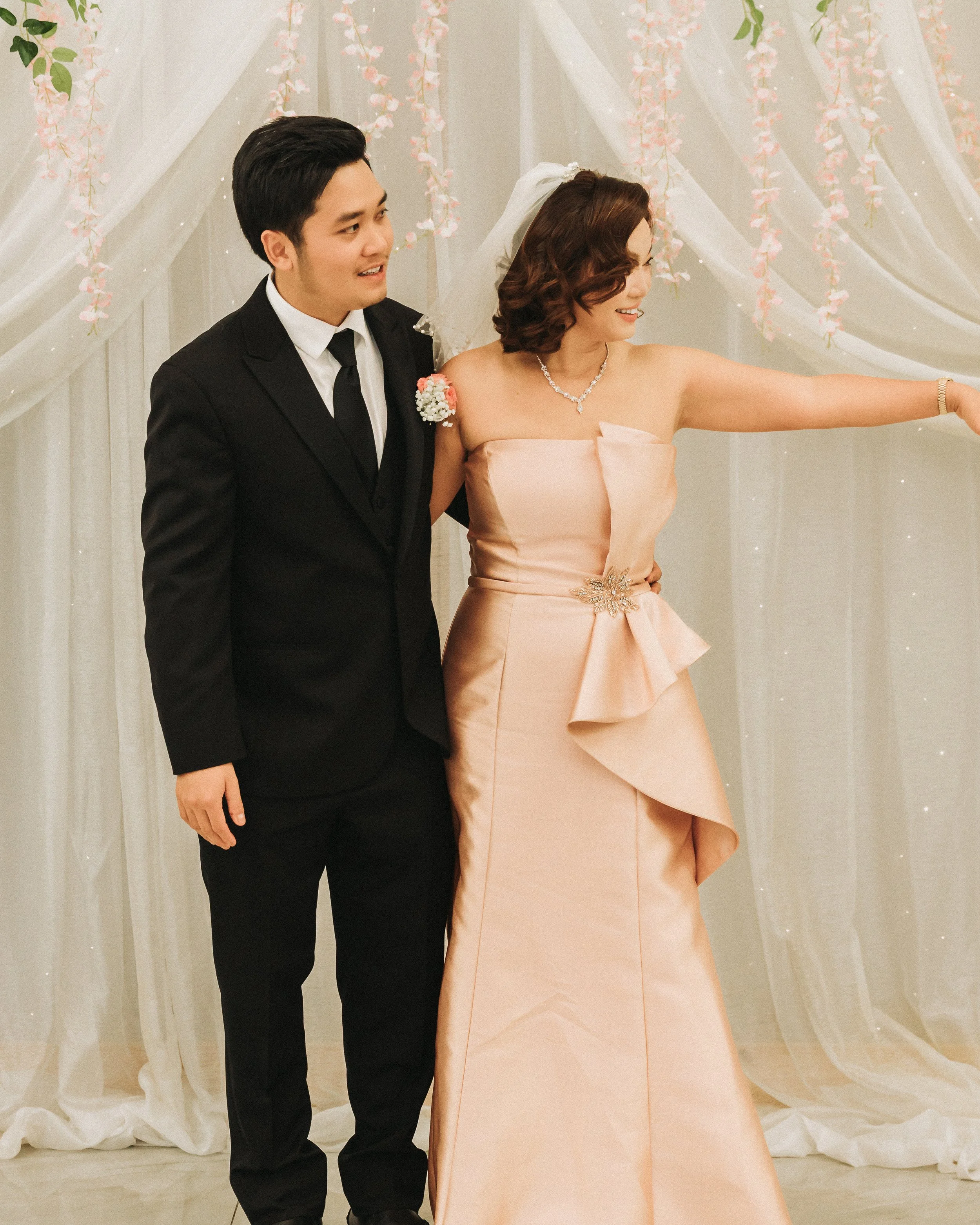 A young man in a black suit and tie standing next to a woman in a peach-colored strapless dress, who is pointing at something in the distance. They are in front of a decorative backdrop with hanging pink flowers and white drapes.