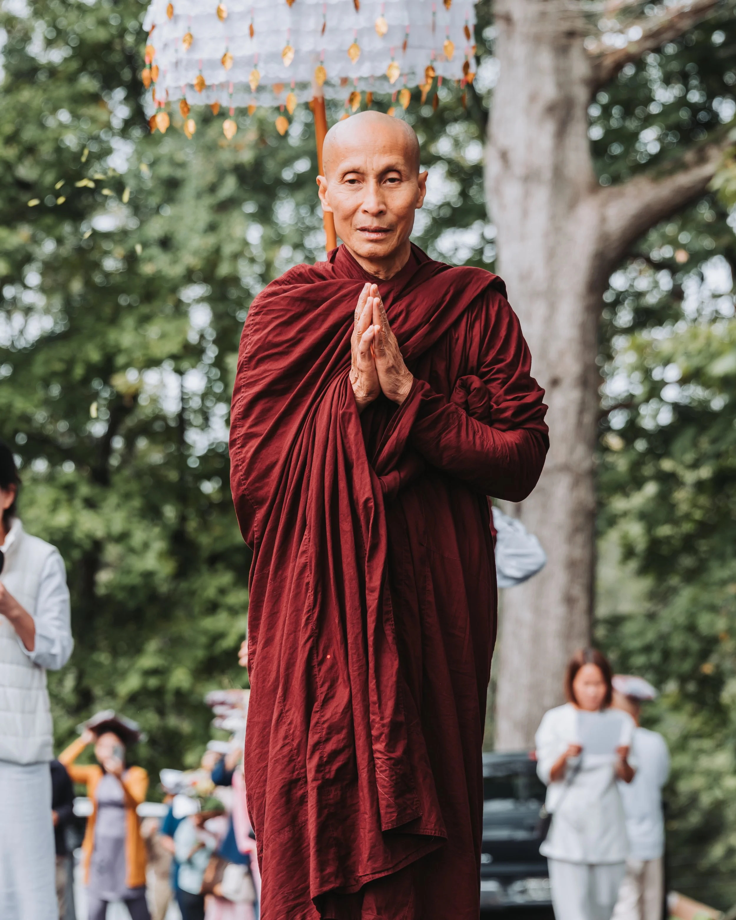 A Buddhist monk dressed in maroon robes standing outdoors in a park with hands pressed together in a prayer gesture, surrounded by others and trees.